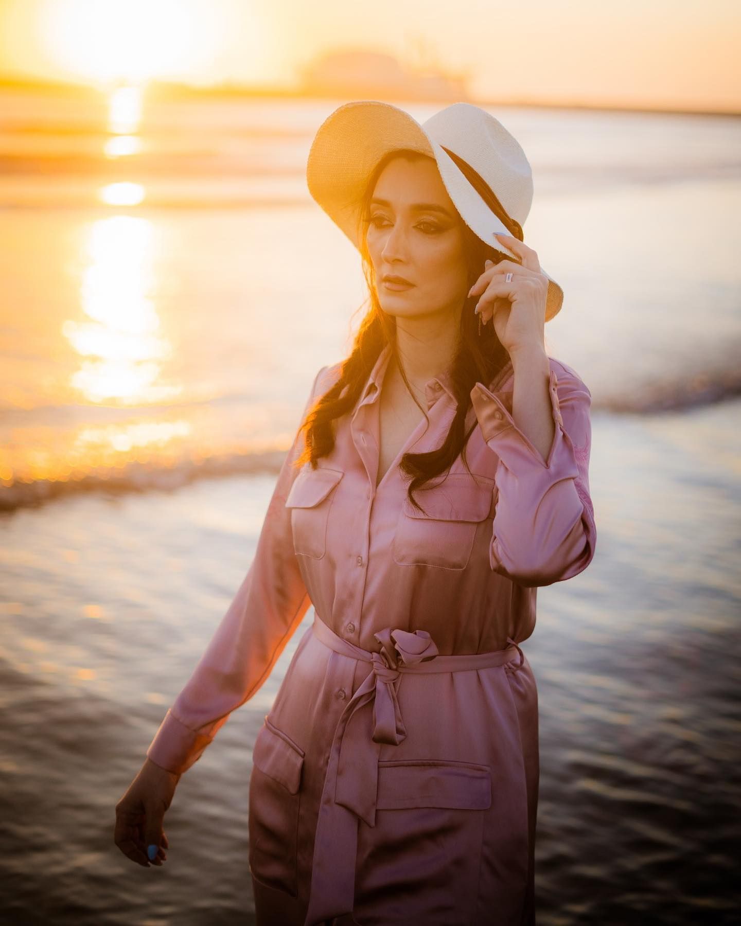 Elegant woman in pink belted dress and white sun hat walking on beach at golden sunset.