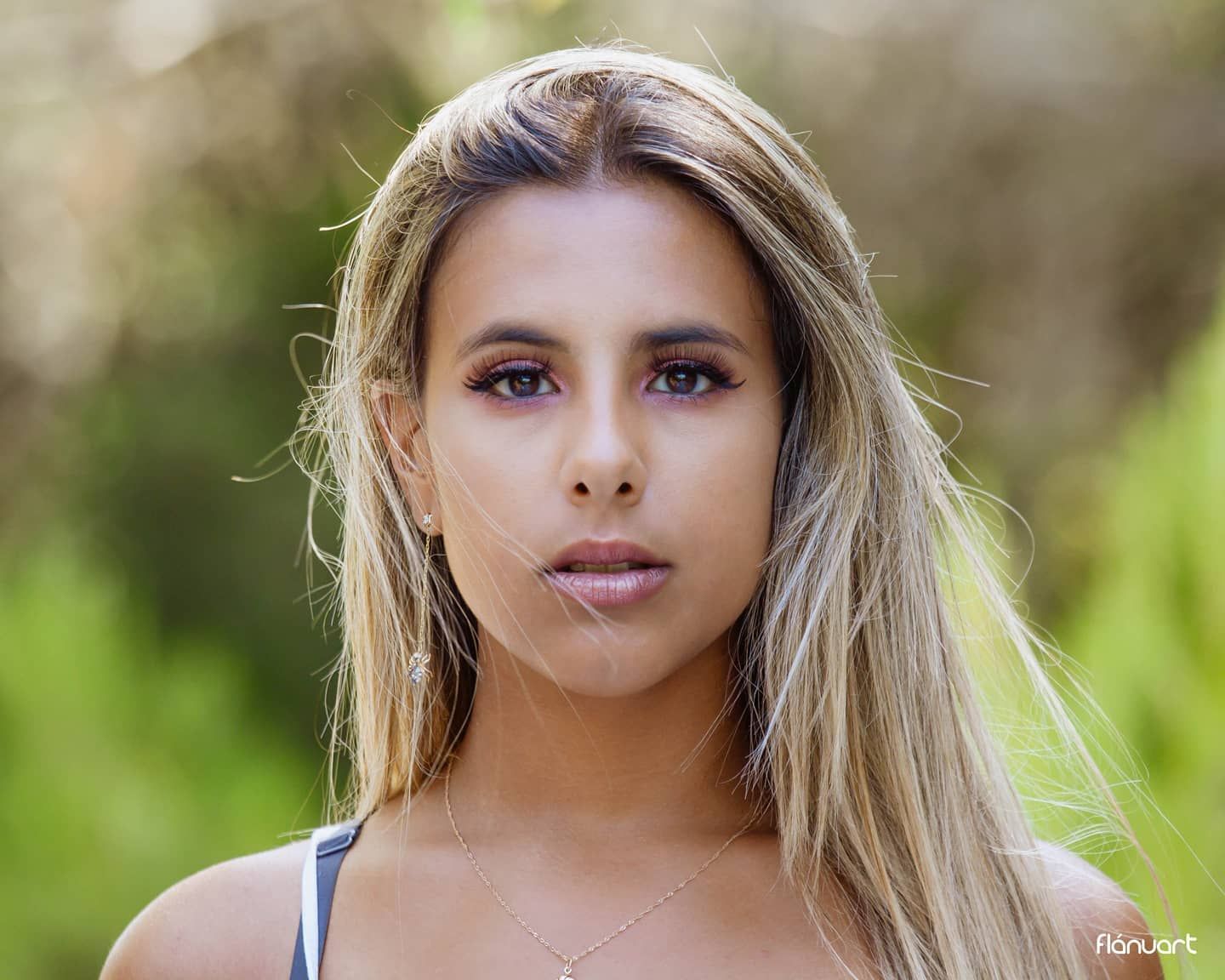 Close-up portrait of blonde woman with natural makeup in an outdoor green setting.