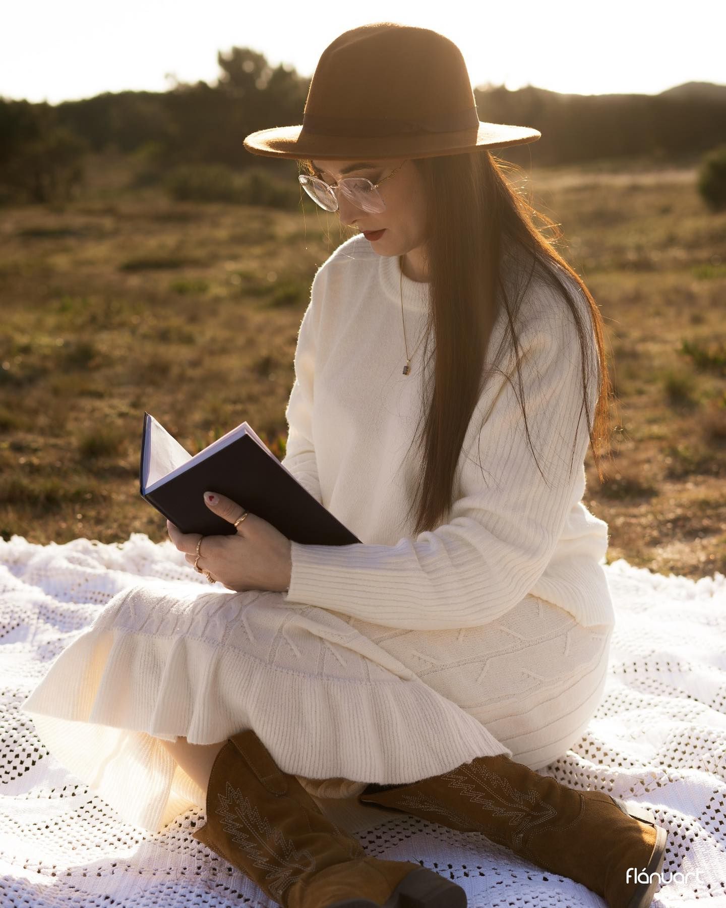 Woman in cream knit dress and wide-brim hat reading book in sunny field.