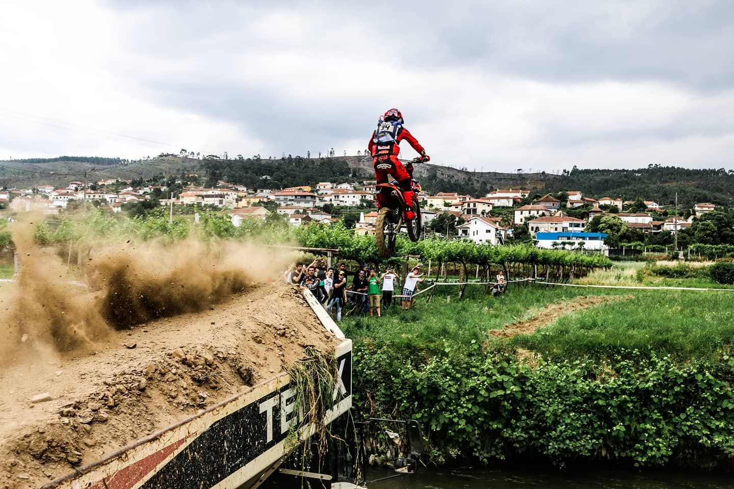 Motocross rider launching off dirt jump with spectators and village in background.