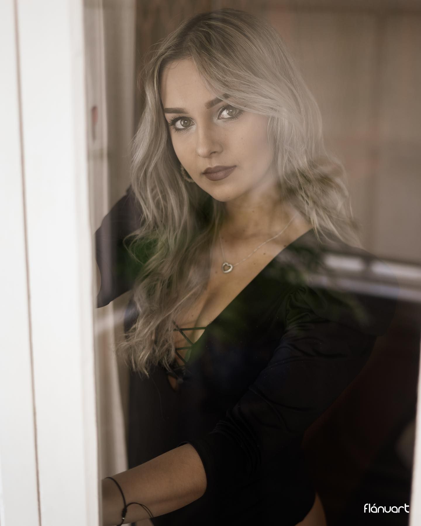 Blonde woman in black V-neck top gazes through glass door wearing gold necklace.
