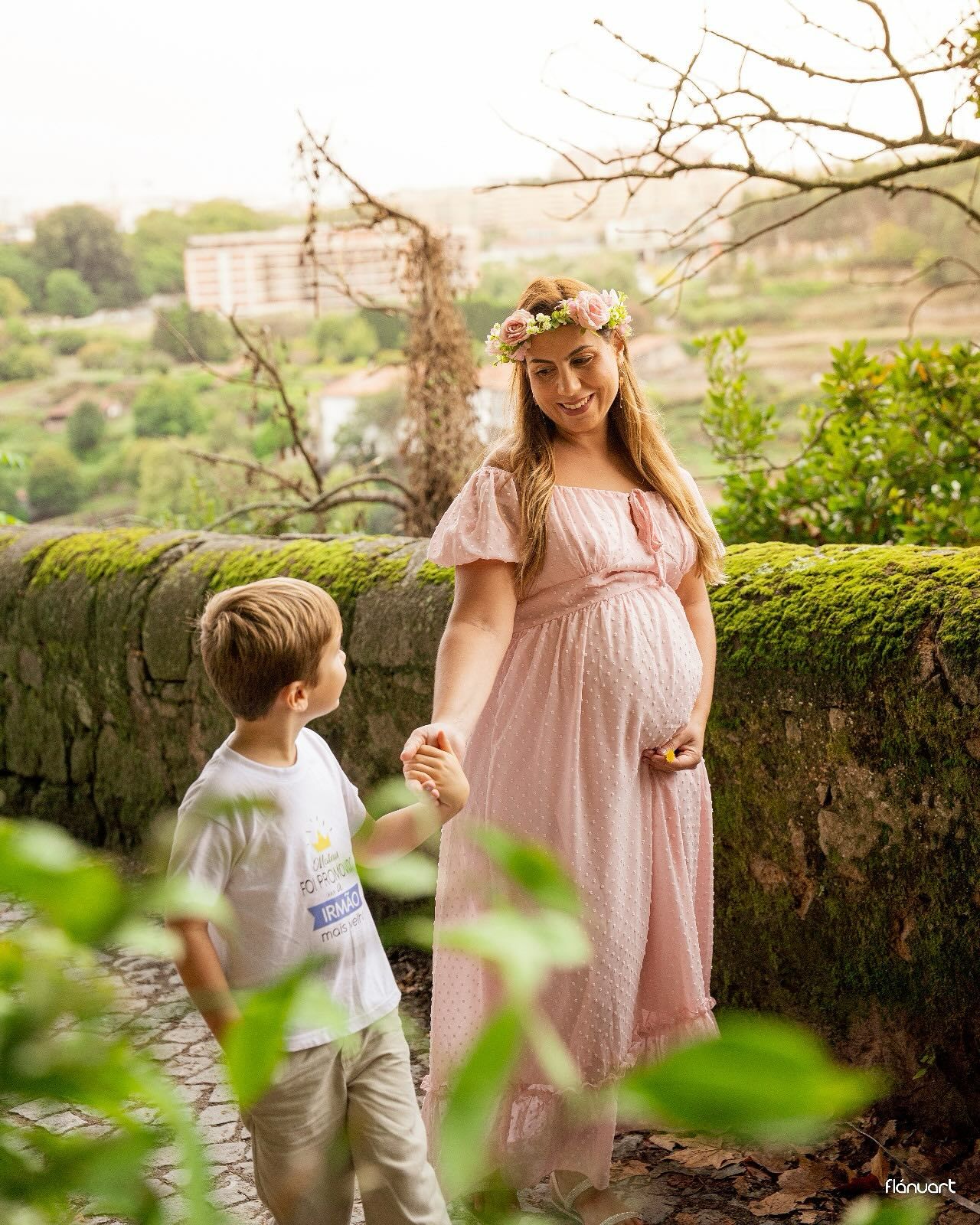 Pregnant woman in pink dress with flower crown holding hands with young boy in garden.