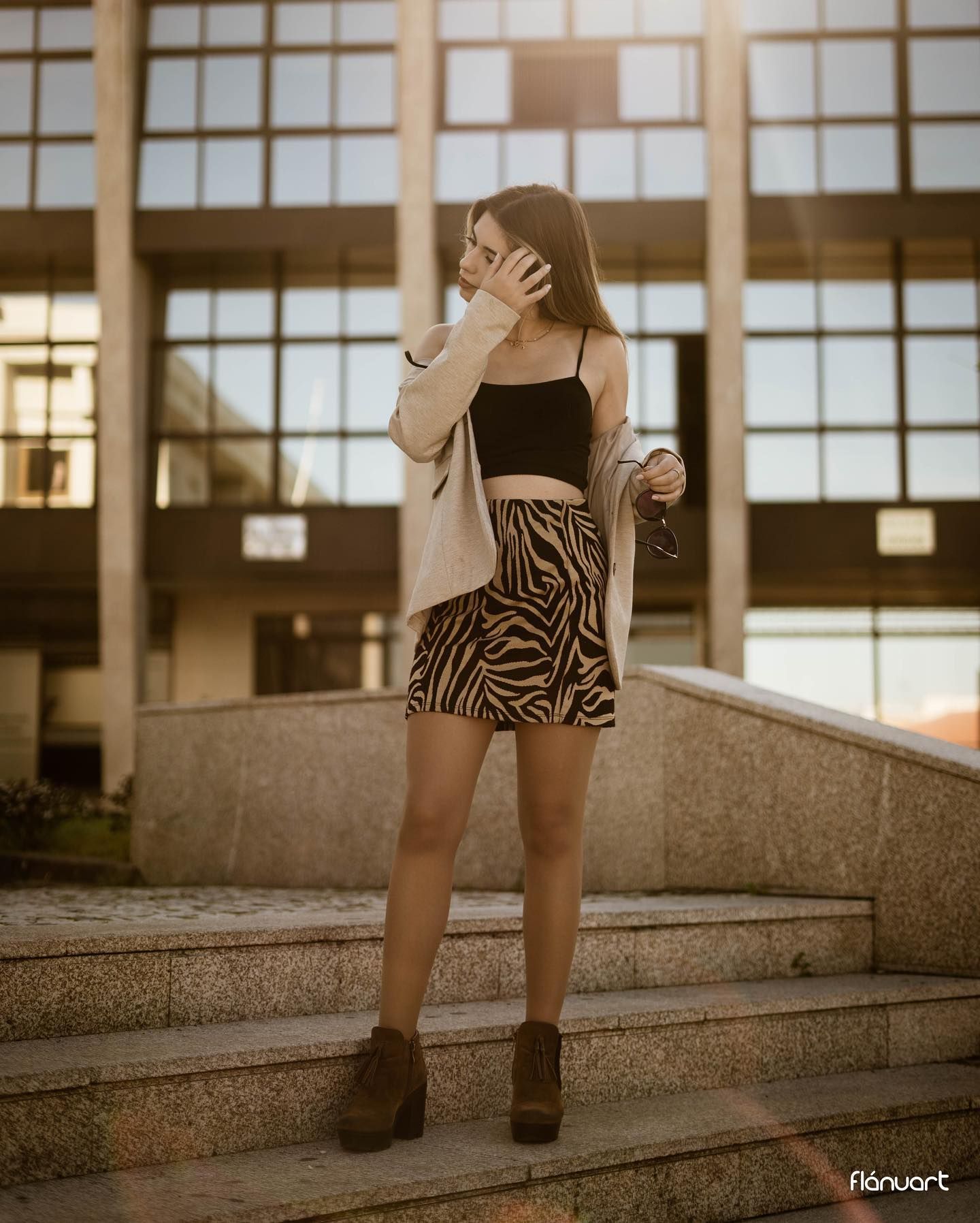 Young woman in zebra print mini skirt, black crop top, and beige cardigan posing on outdoor steps at golden hour.