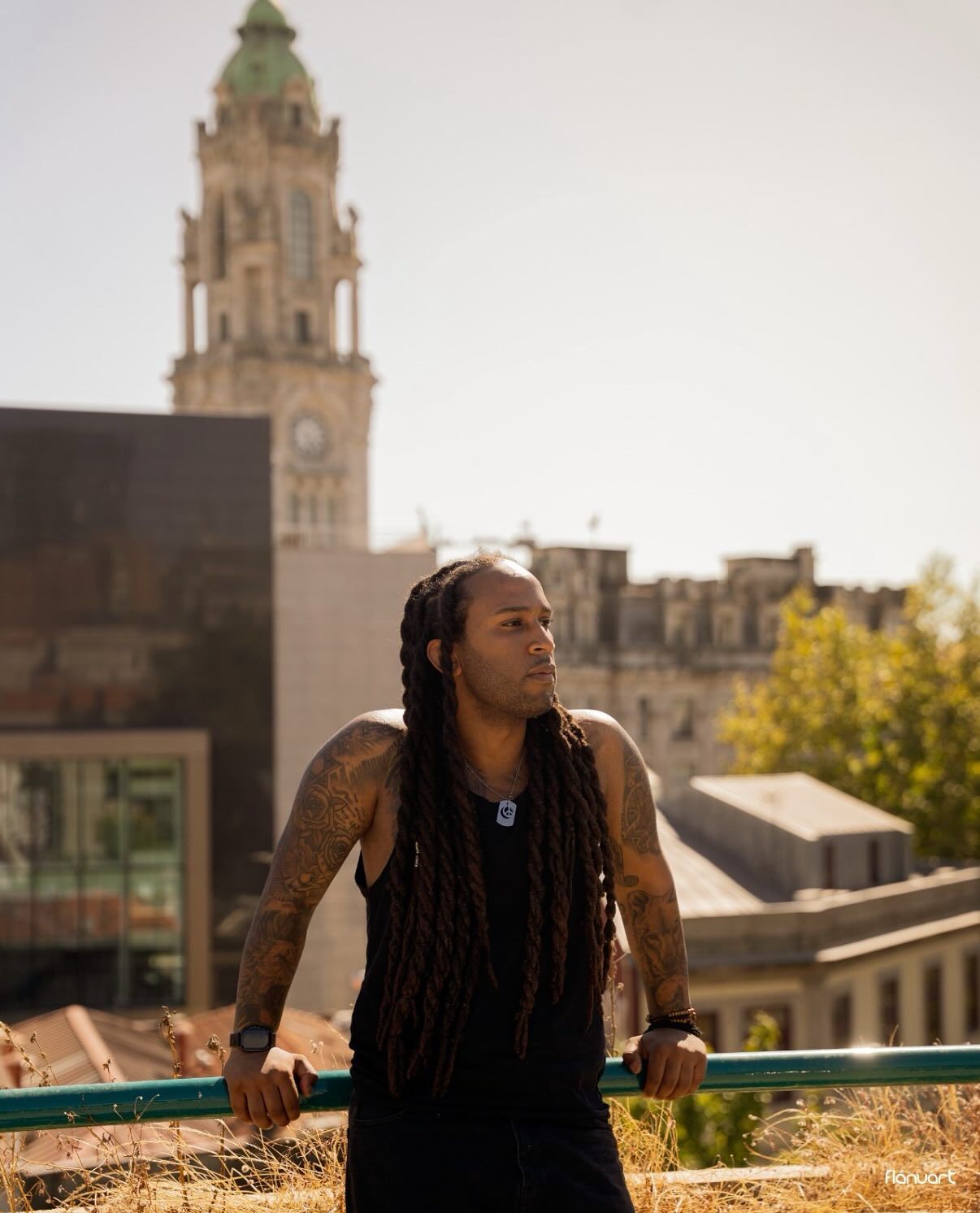 Tattooed man with long dreadlocks leaning on a railing with a historic clock tower in the background.