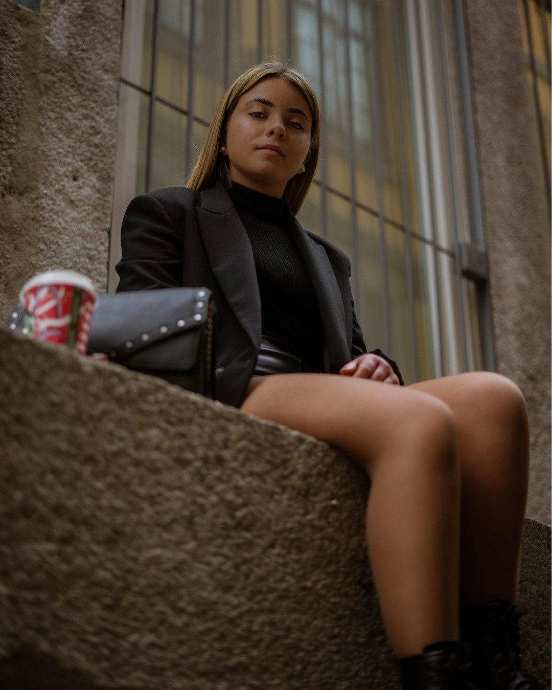 Young woman in black blazer and turtleneck sits on ledge with studded bag and coffee.