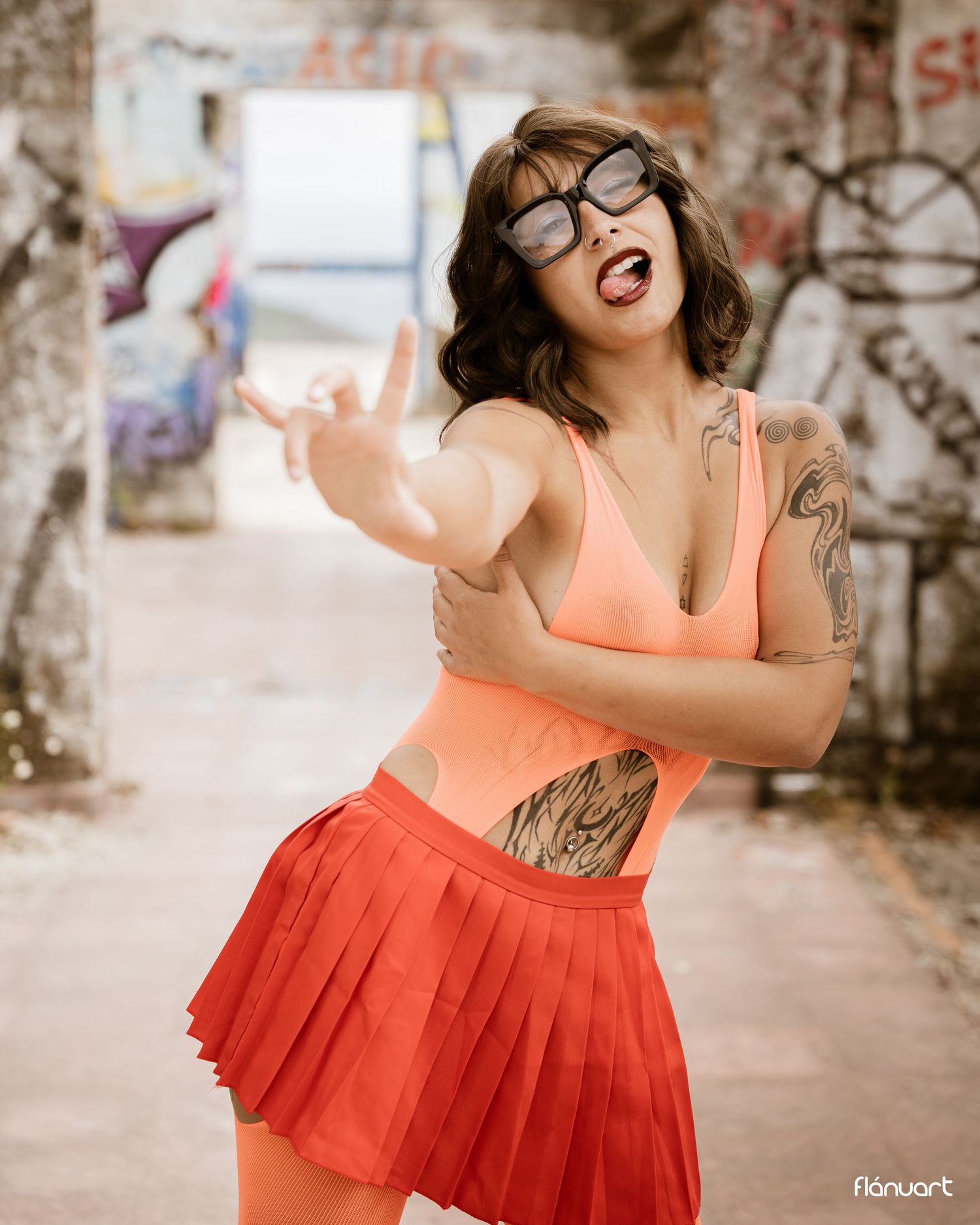 Woman in orange bikini top and red pleated skirt with tattoos and sunglasses posing near graffiti wall.