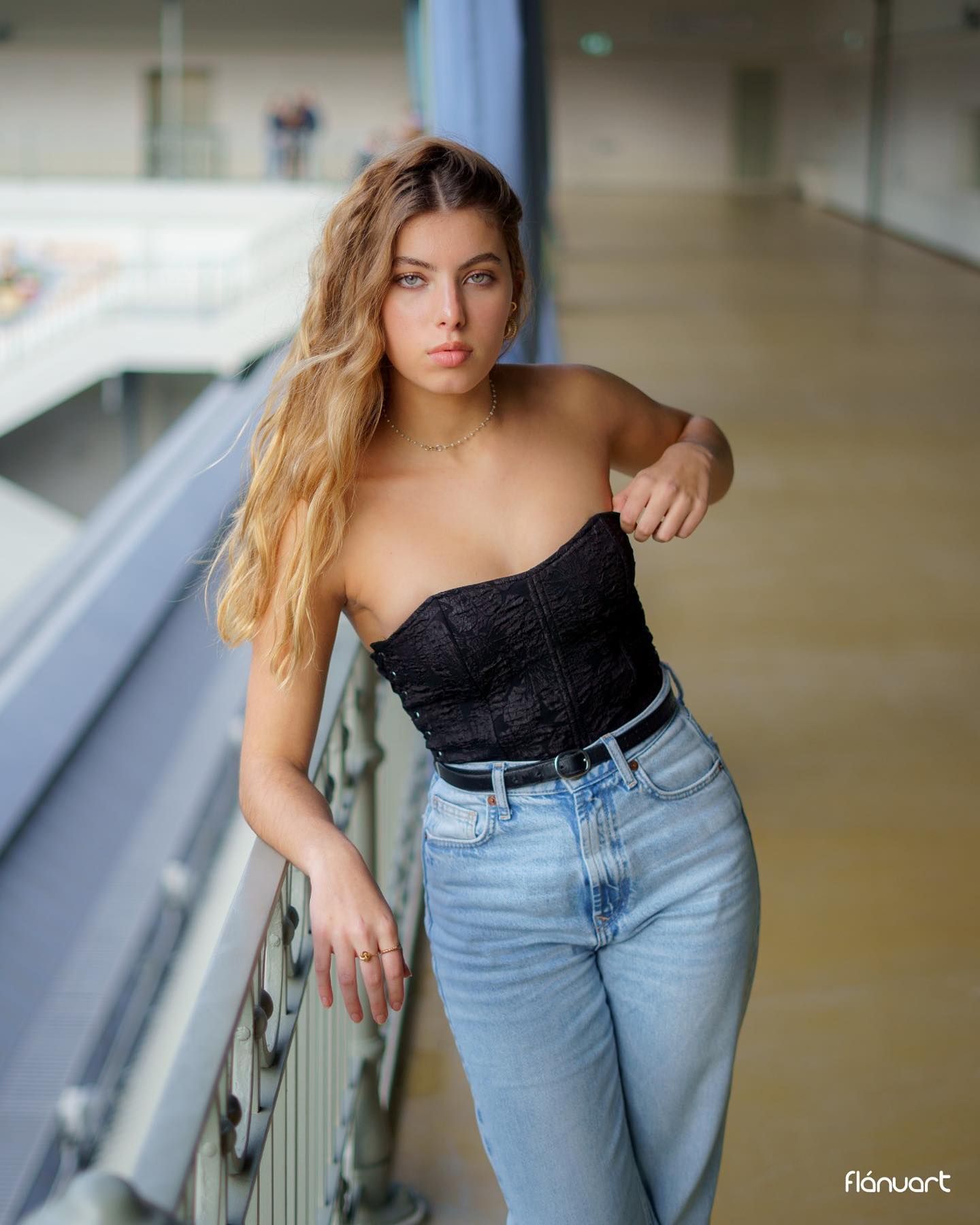 Woman in black corset top and jeans leaning on railing in modern building interior.