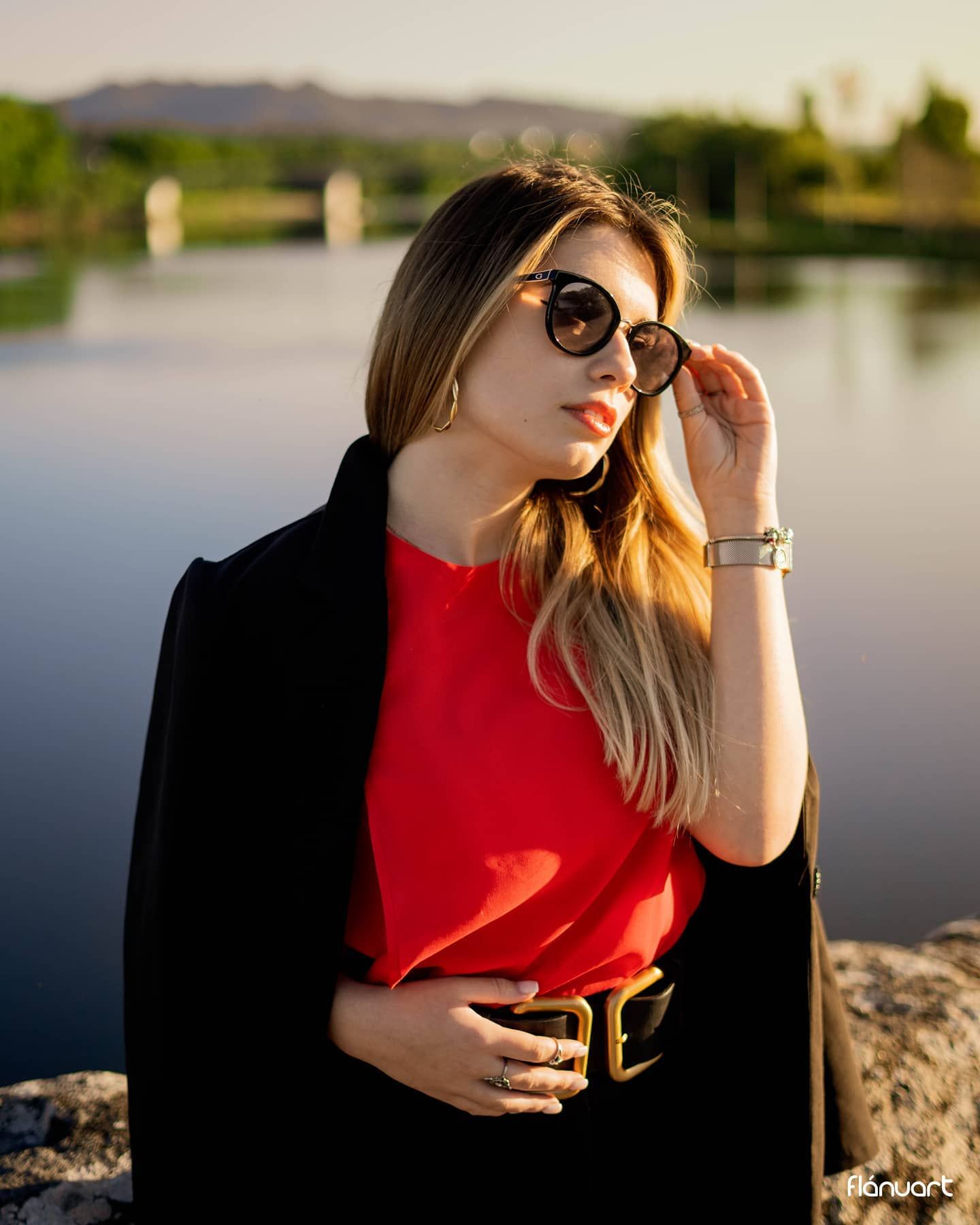 Stylish woman in red top and black coat adjusts sunglasses near a scenic waterfront.