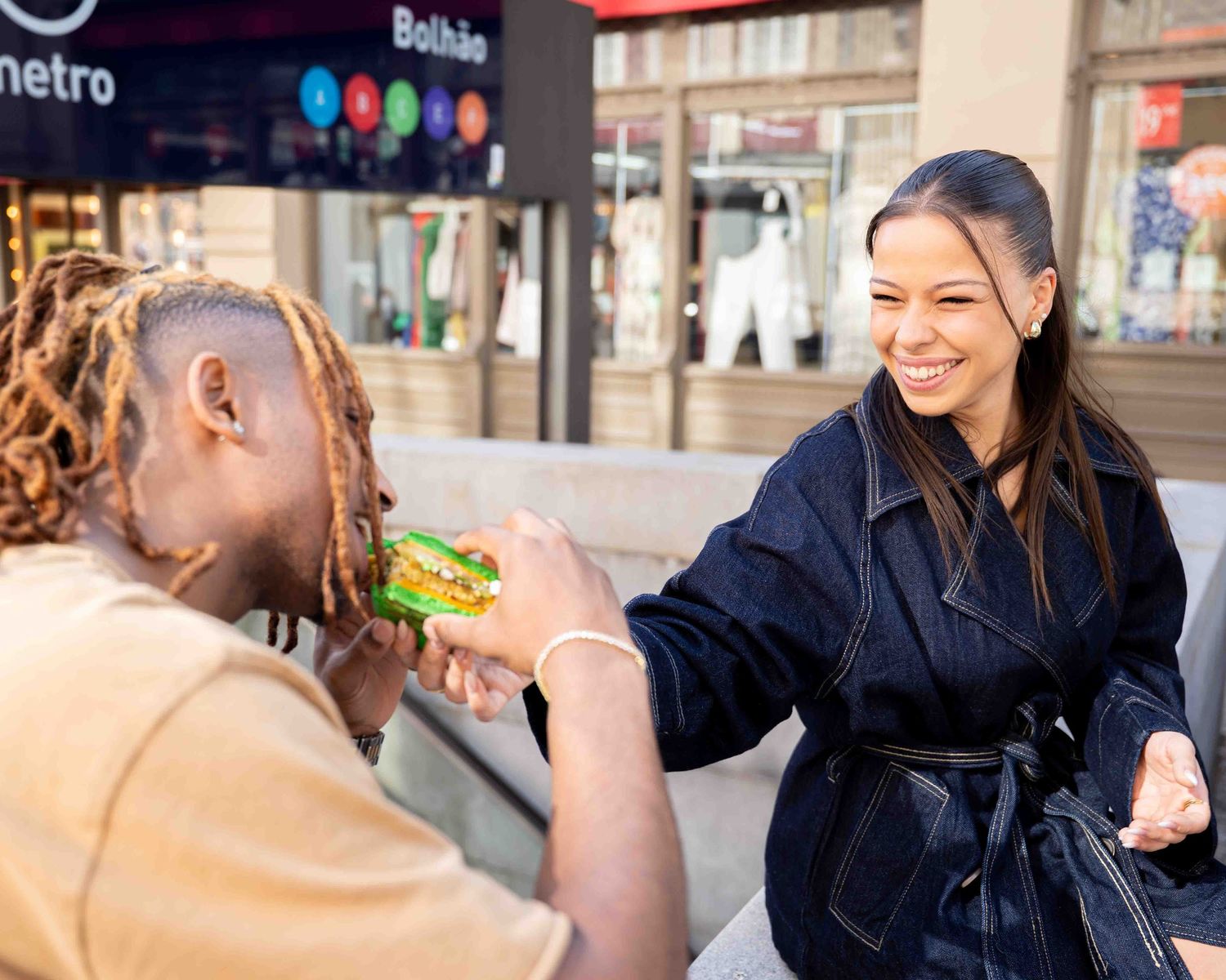 Two friends sharing food and laughing outdoors near a metro station on a sunny day.