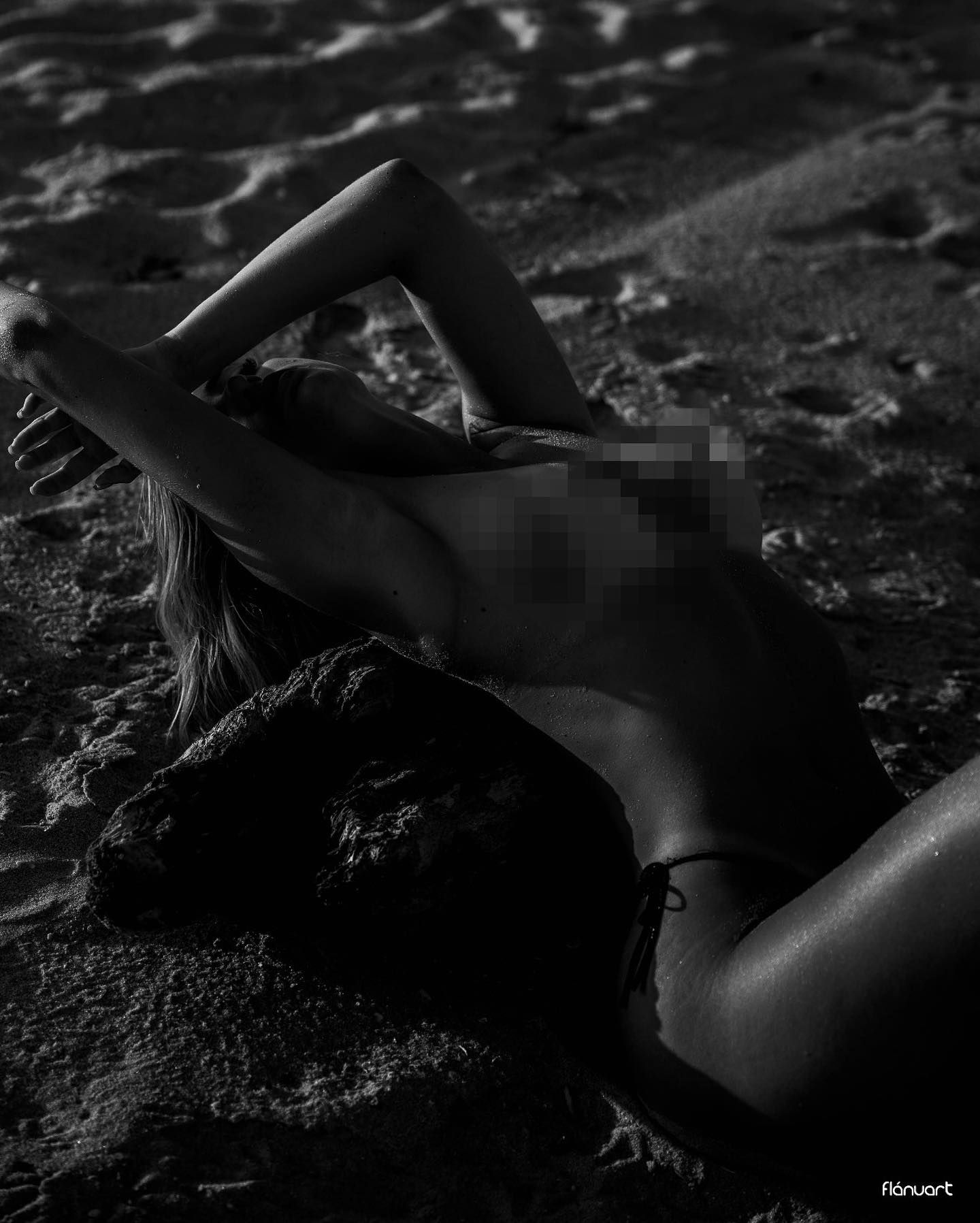 Black and white photo of a blonde woman lying on sandy beach in bikini bottoms, posed artistically.