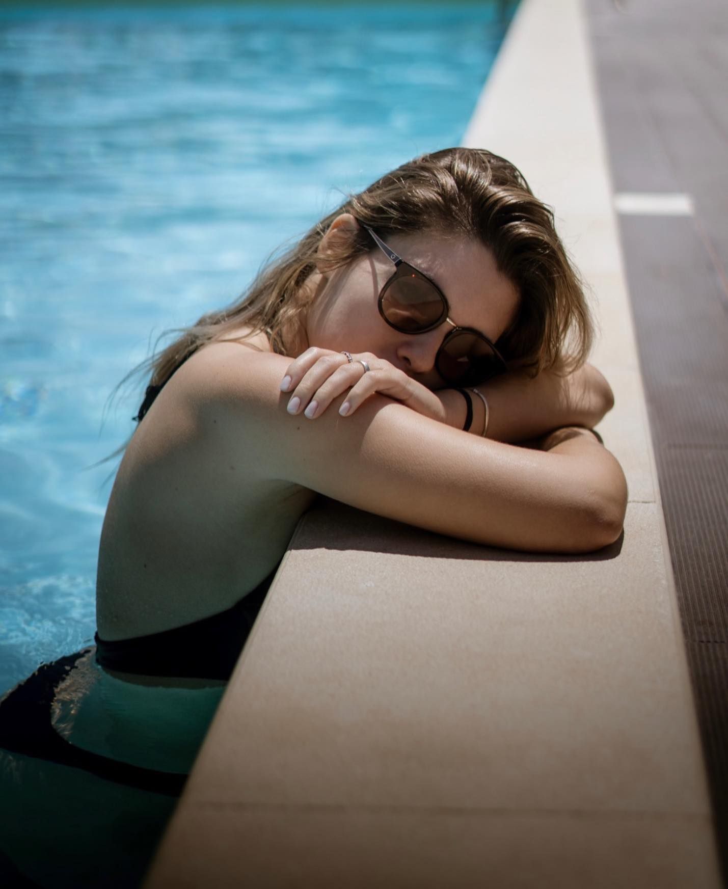 Woman in green swimsuit resting on pool edge with sunglasses and wet hair in natural light.