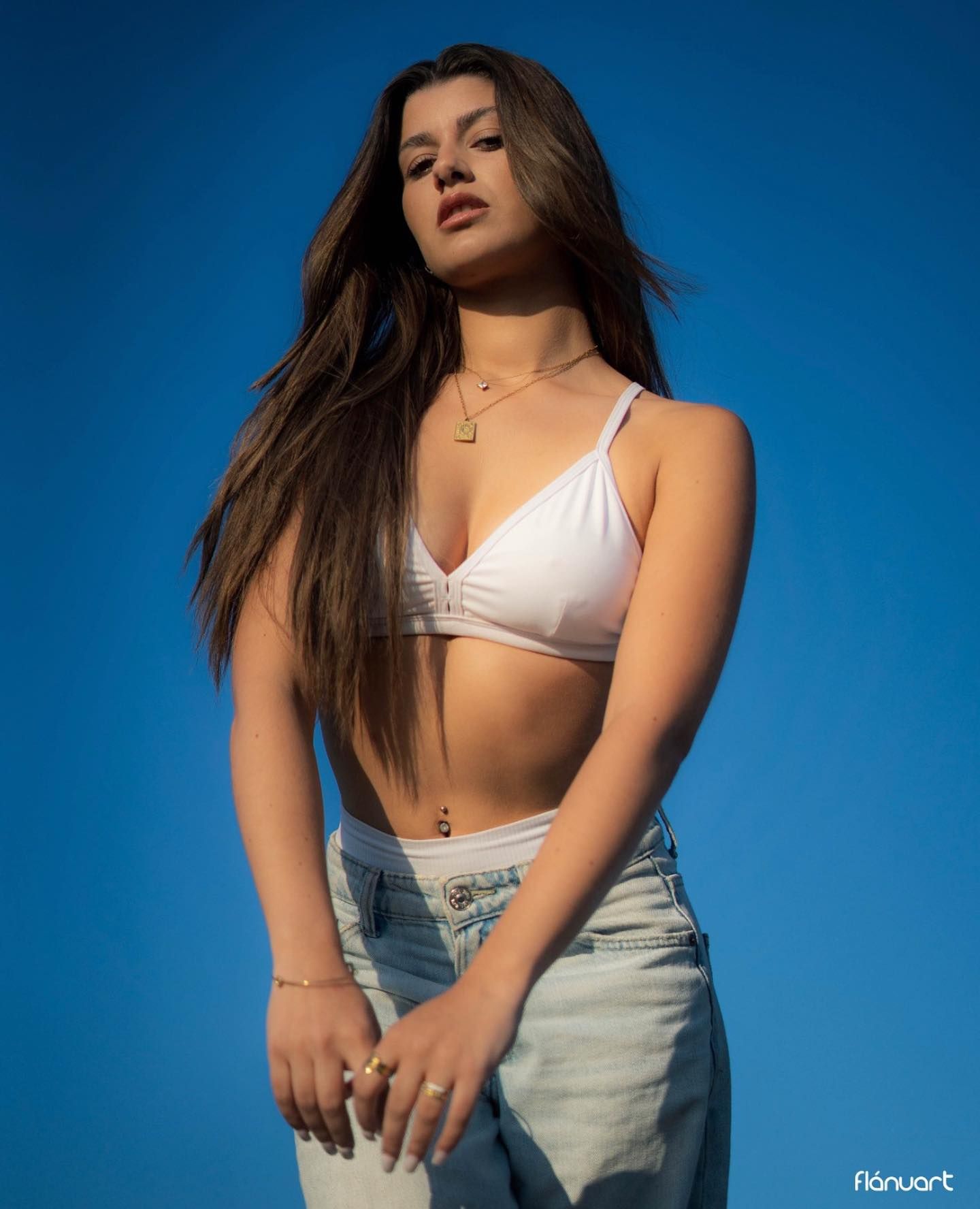 Young woman in white bralette and jeans posing confidently against bright blue background.