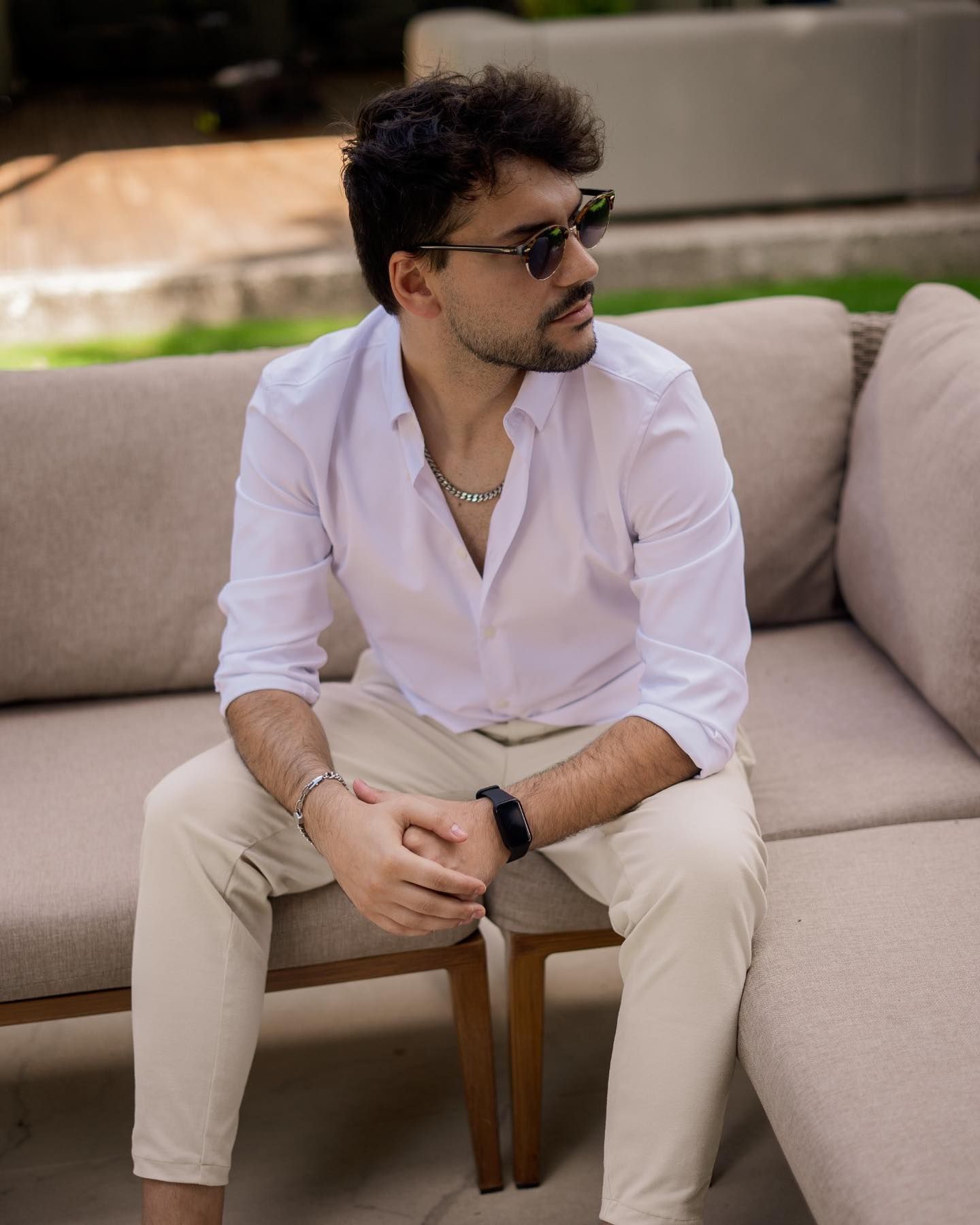 Stylish man in white shirt and beige pants sitting on outdoor sofa wearing sunglasses.