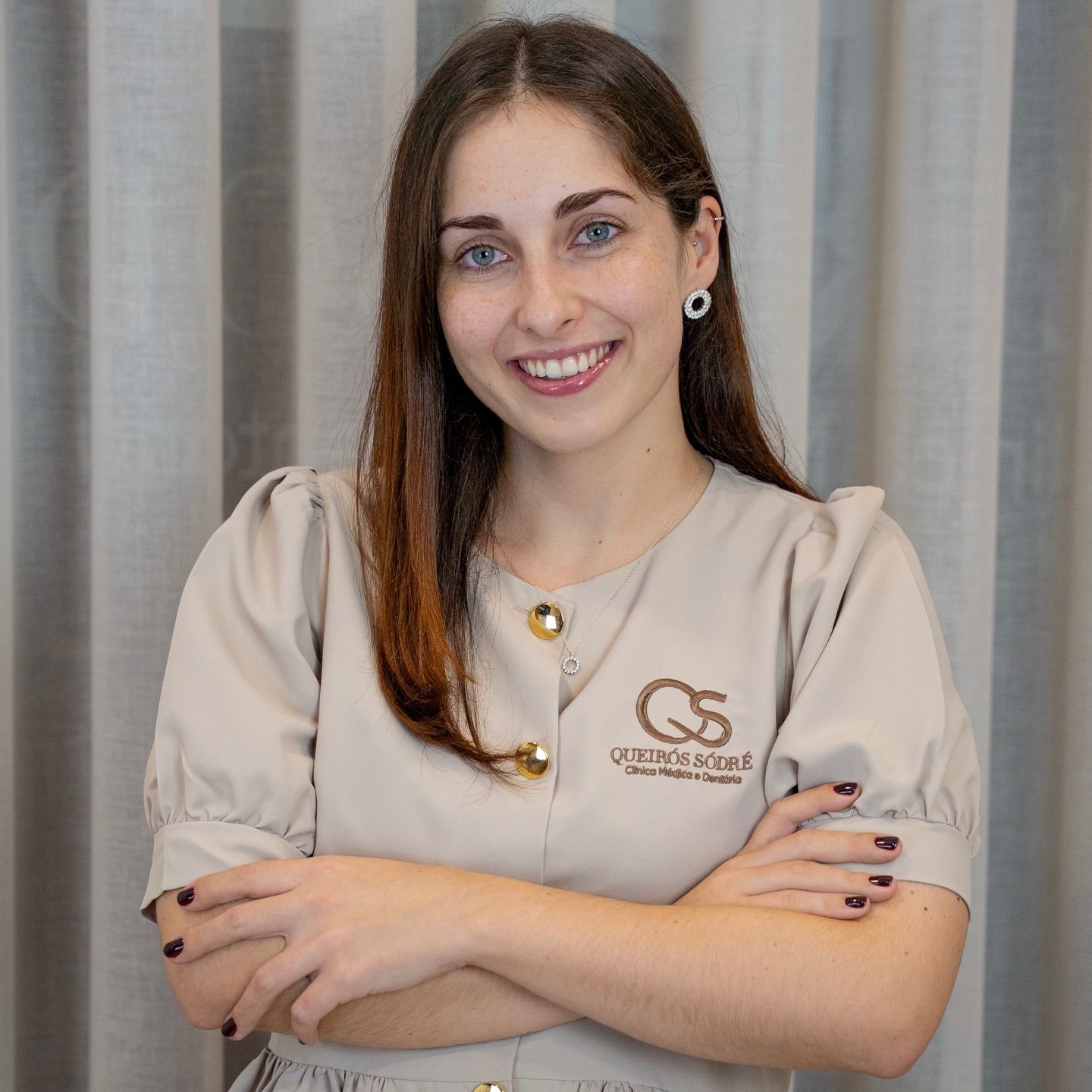 Young female medical professional in beige scrubs smiling with arms crossed in a clinic setting.