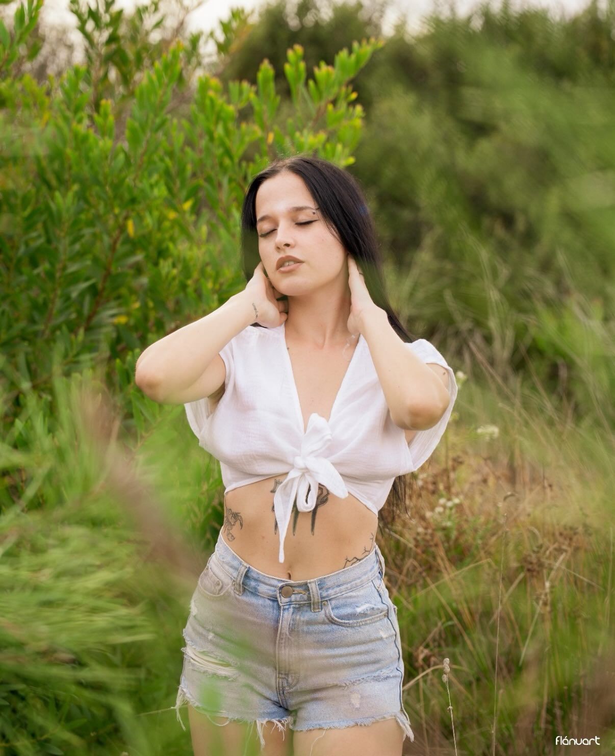 Young woman in tied white crop top and denim shorts posing among green bushes outdoors.