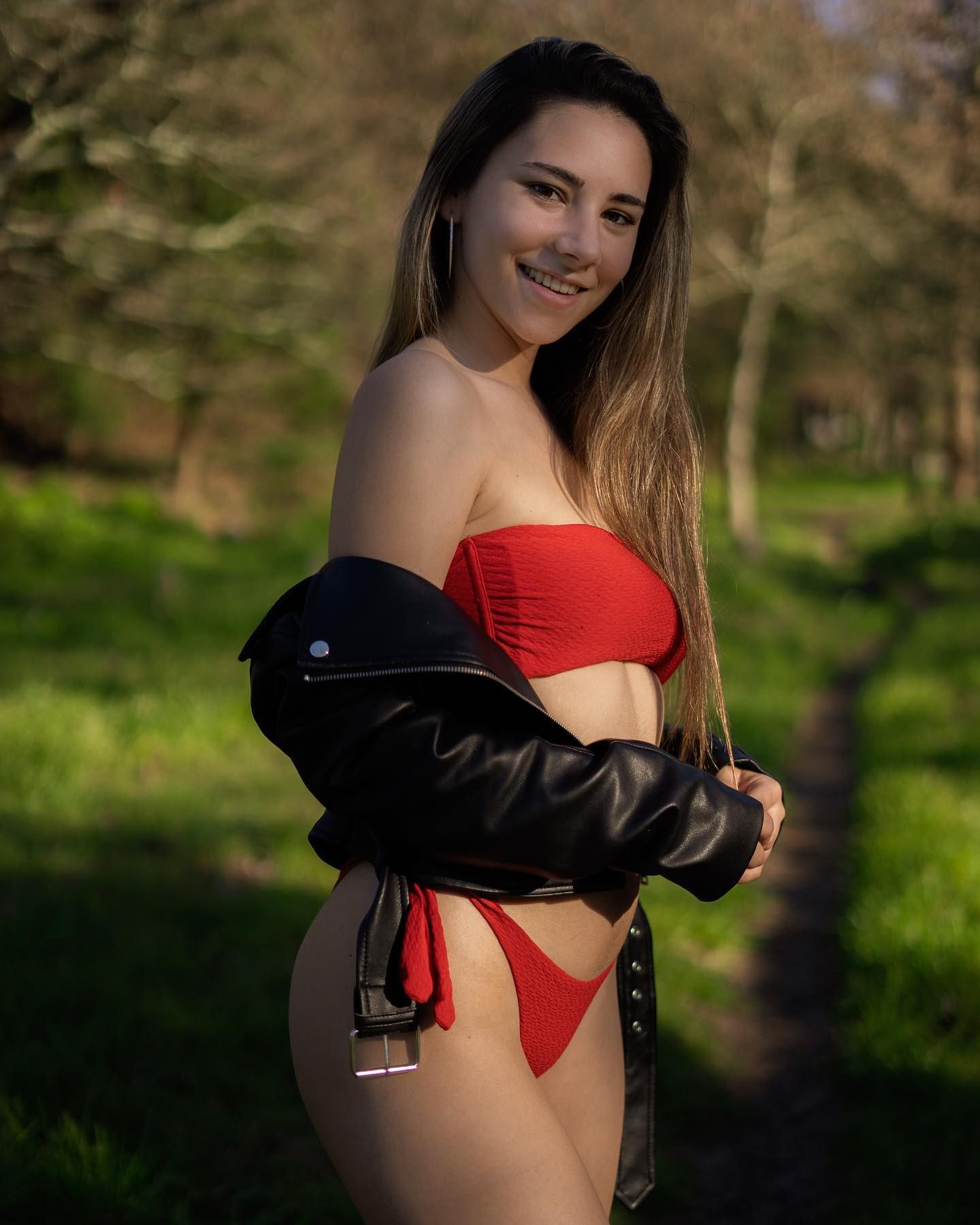 Smiling woman in red bikini and black leather jacket posing in sunny park.