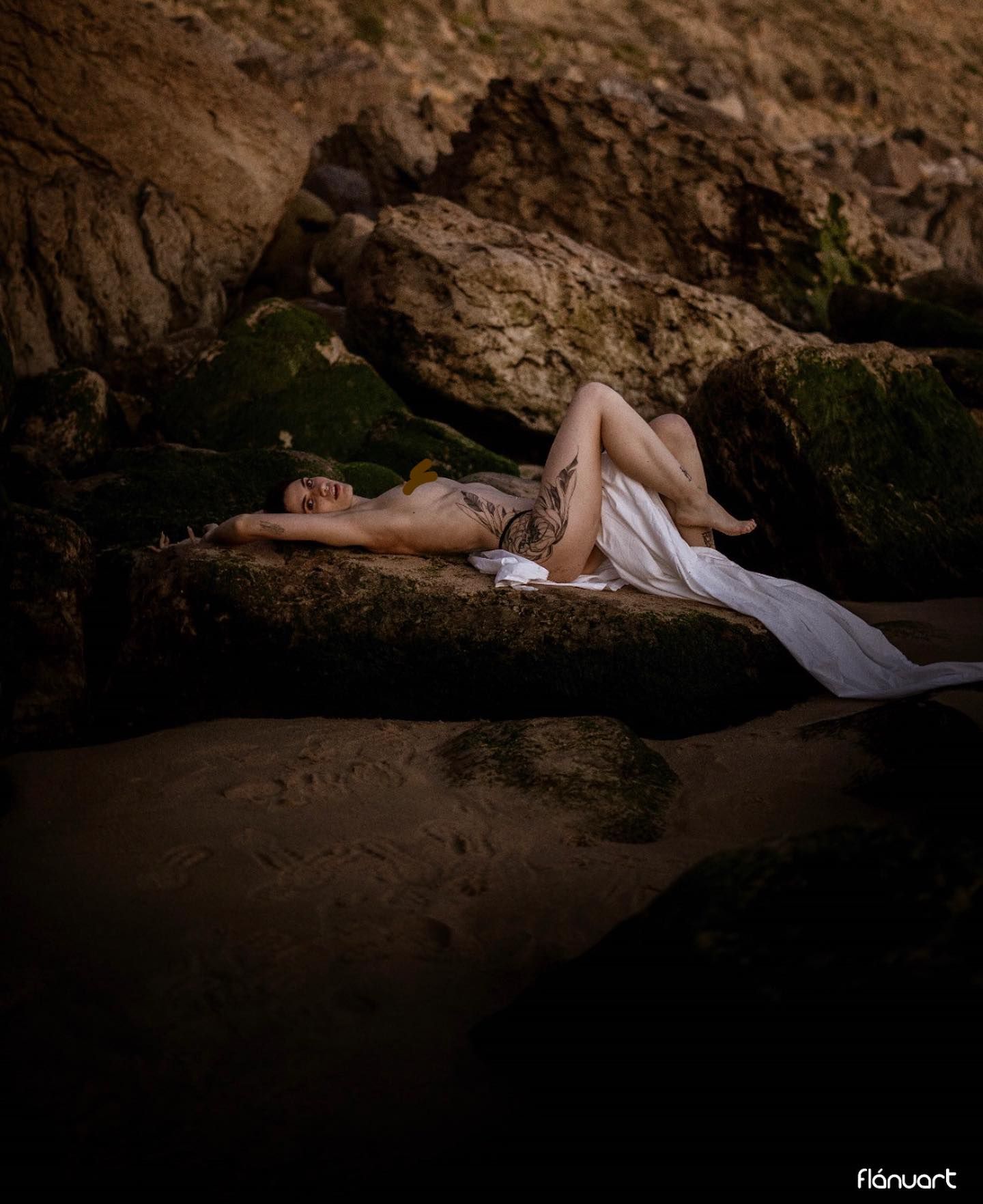 Woman in white draped fabric lying on coastal rocks near dark water at dusk.