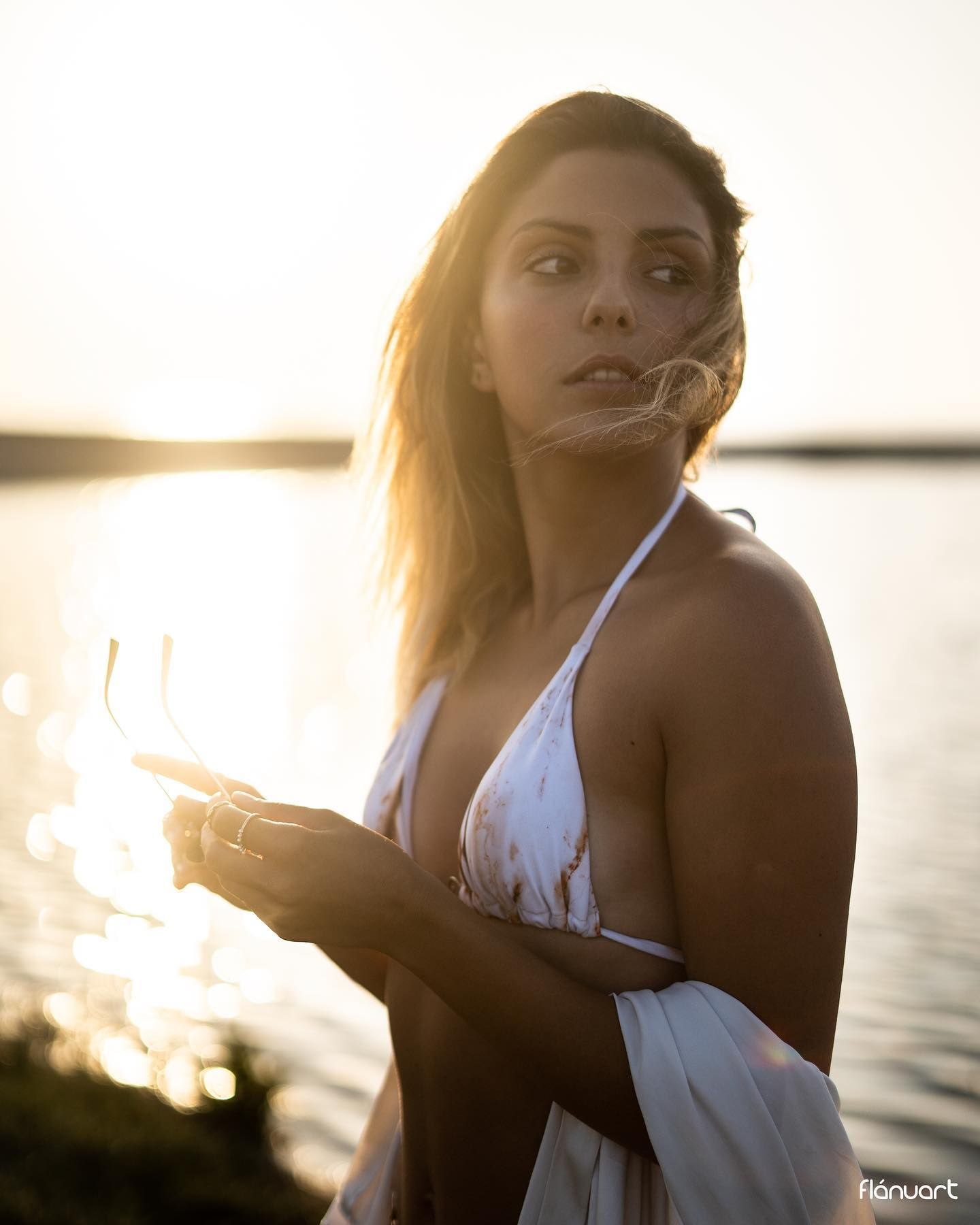 Woman in white bikini holding sunglasses by a lake during a stunning golden sunset.