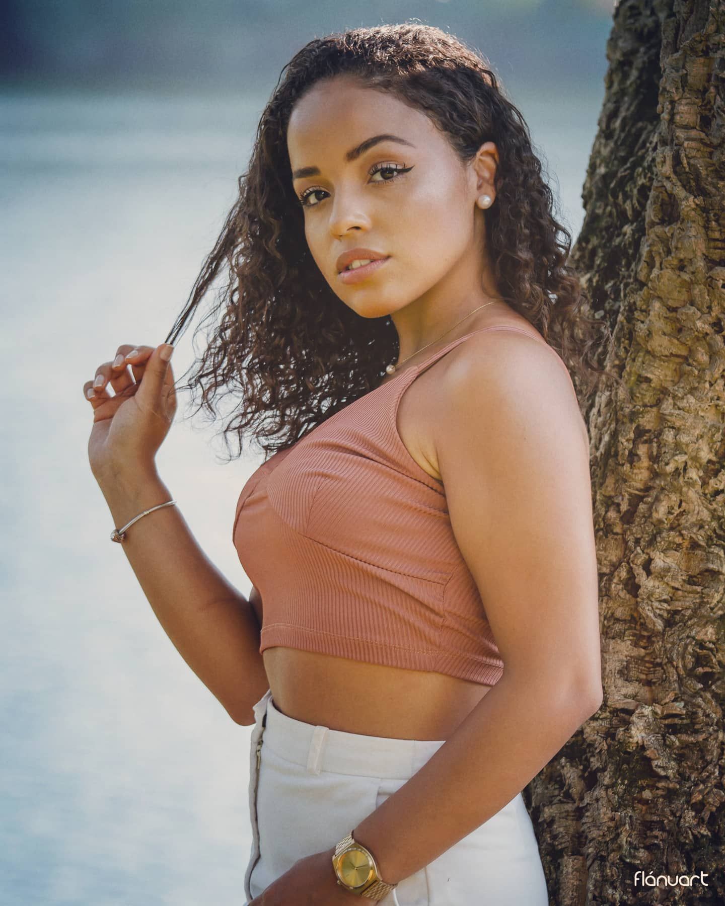 Young curly-haired woman in pink crop top and white skirt leaning against tree by water.
