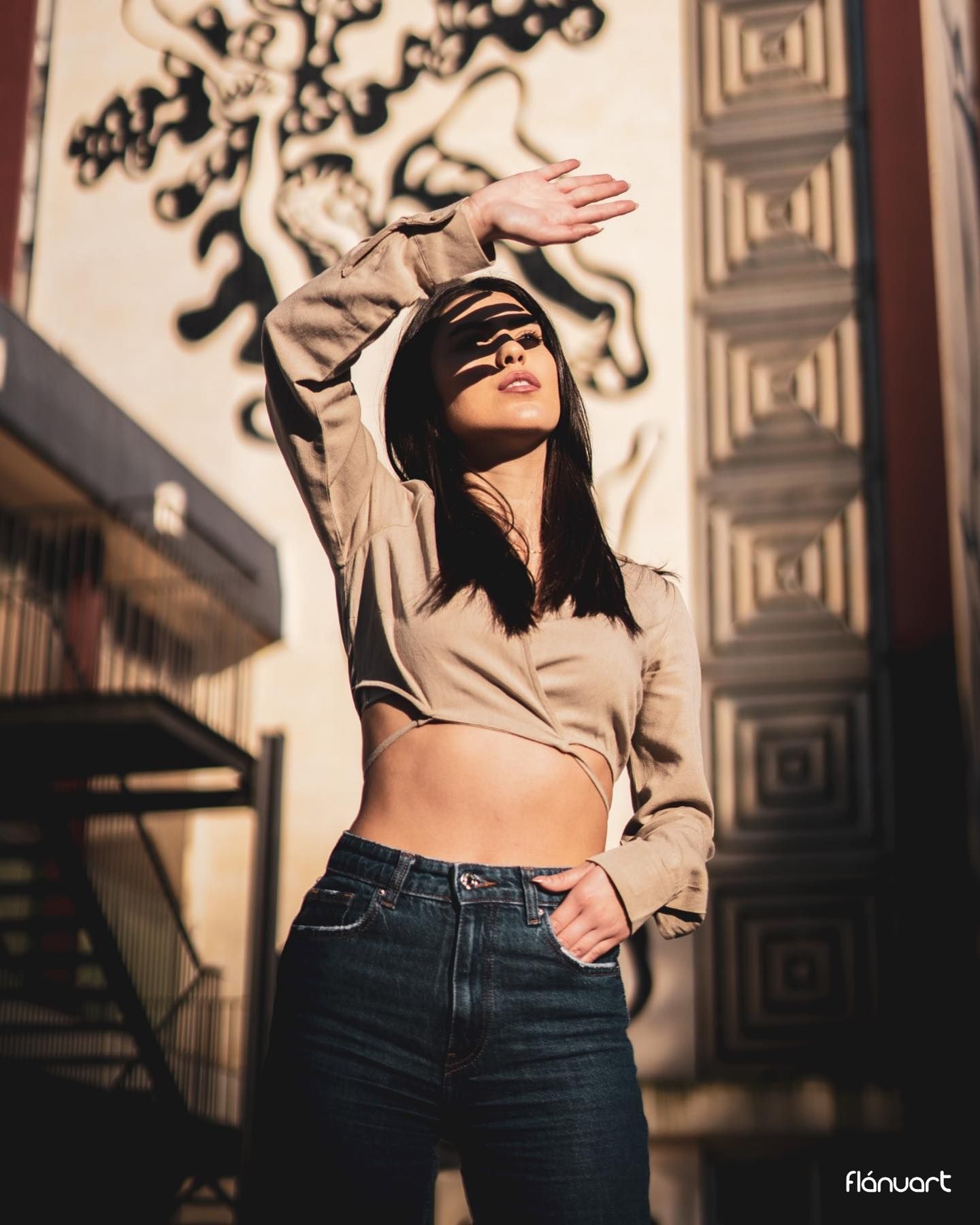Woman in beige crop top and jeans shielding eyes from sun near decorative mural wall.