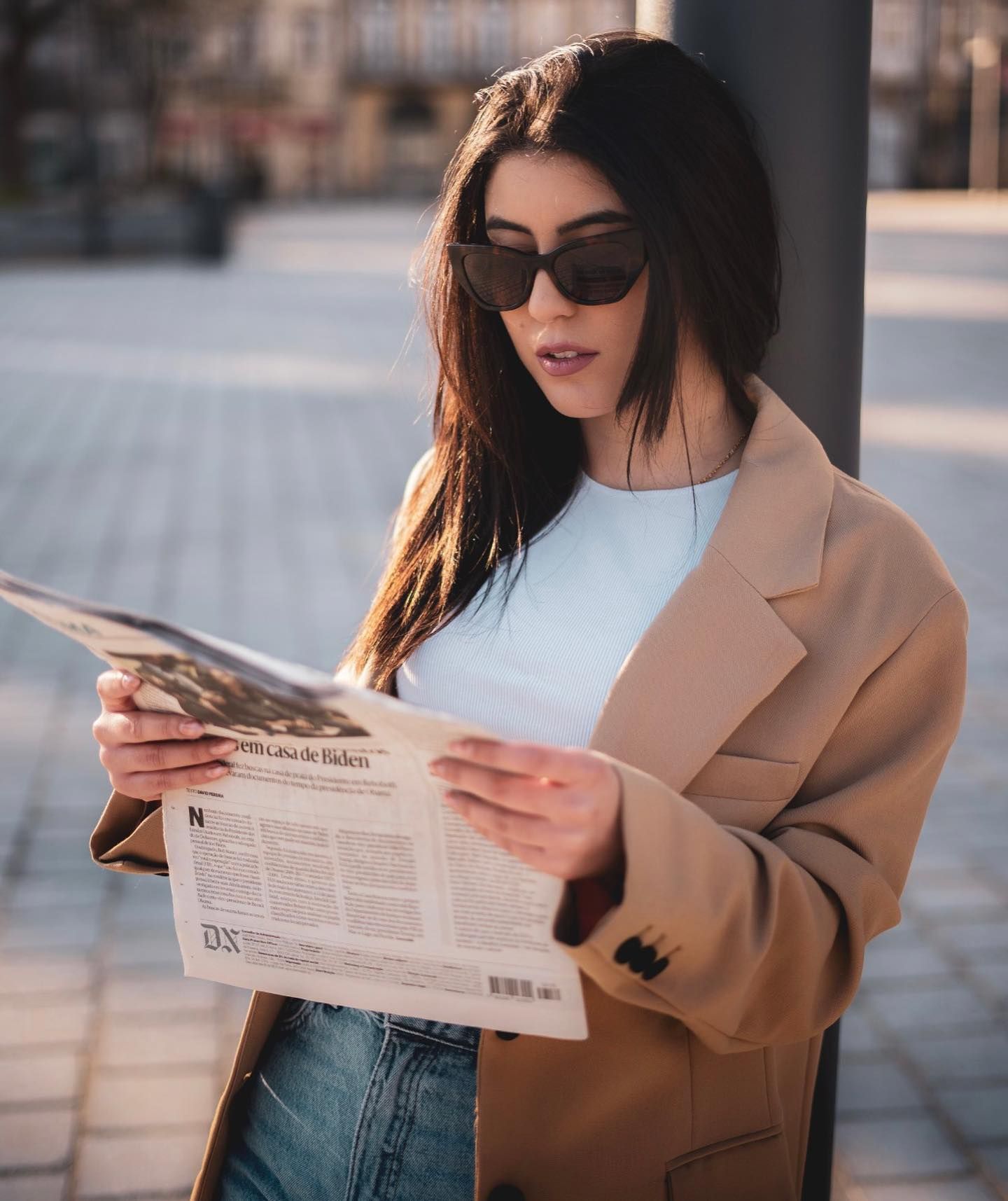 Brunette woman in beige blazer reading newspaper outdoors wearing cat-eye sunglasses.