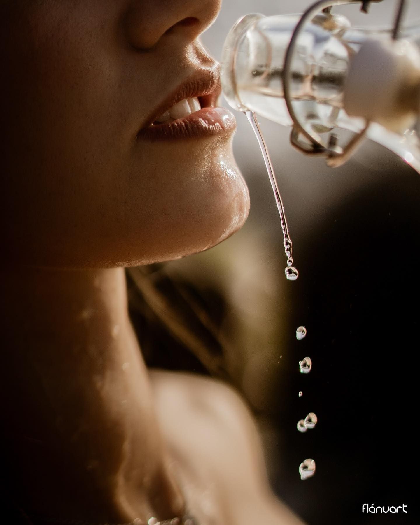 Close-up of woman's lips with water dripping from a glass bottle in warm dramatic lighting.