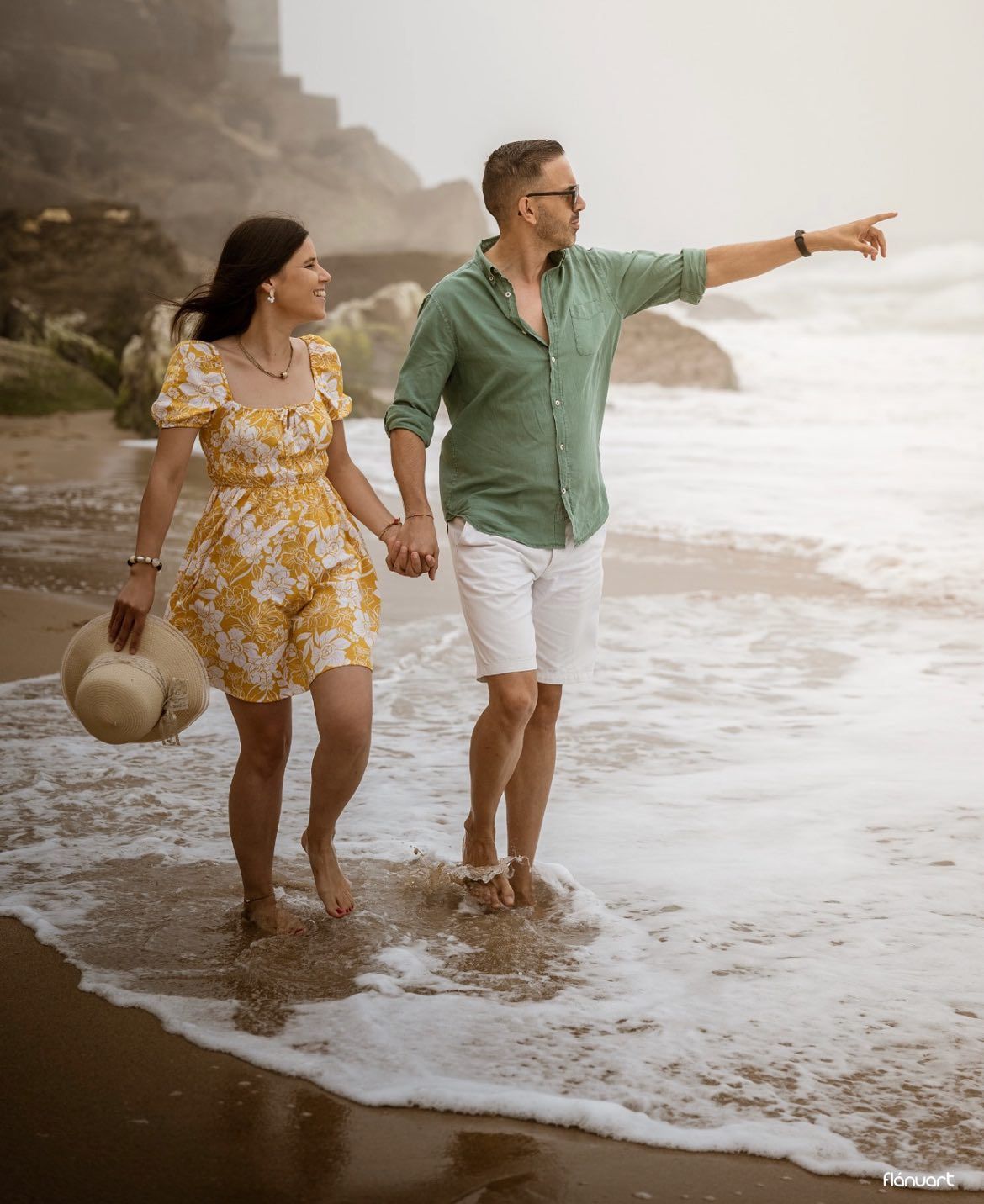 Happy couple walking barefoot on a beach shoreline, man pointing into the distance.