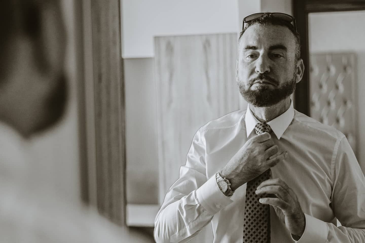 A bearded groom adjusts his tie while looking in a mirror in black and white photo.