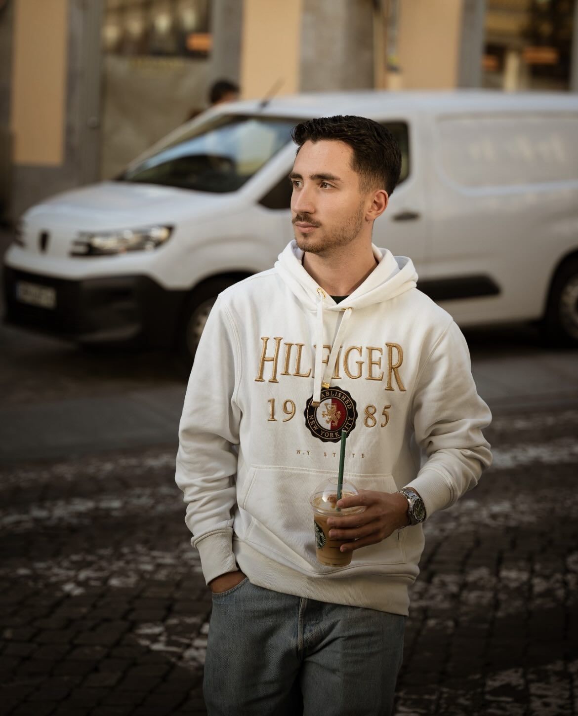 Young man in Tommy Hilfiger hoodie holding iced coffee on a cobblestone city street.