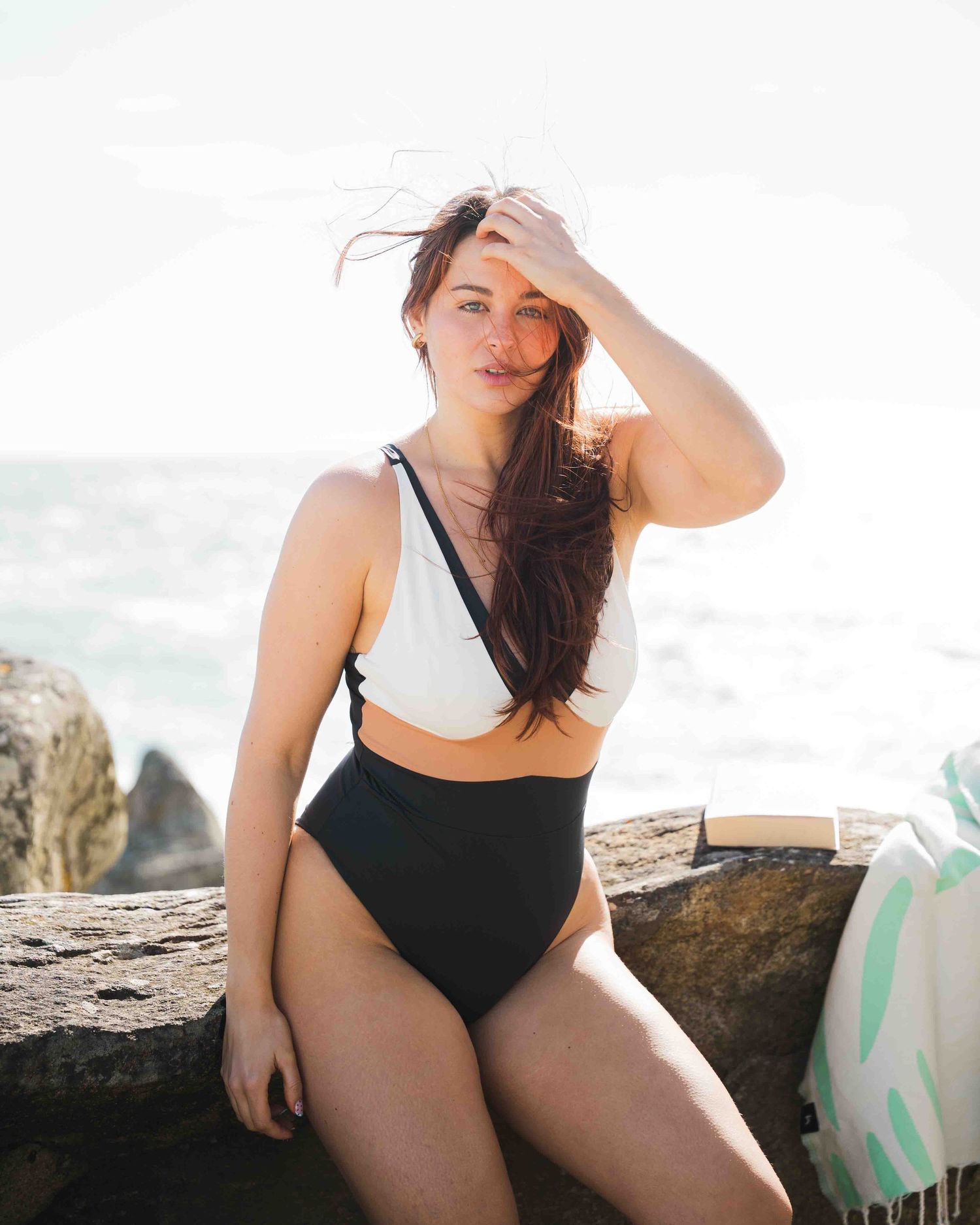 Woman in black and white one-piece swimsuit posing on rocks by the ocean on a sunny day.