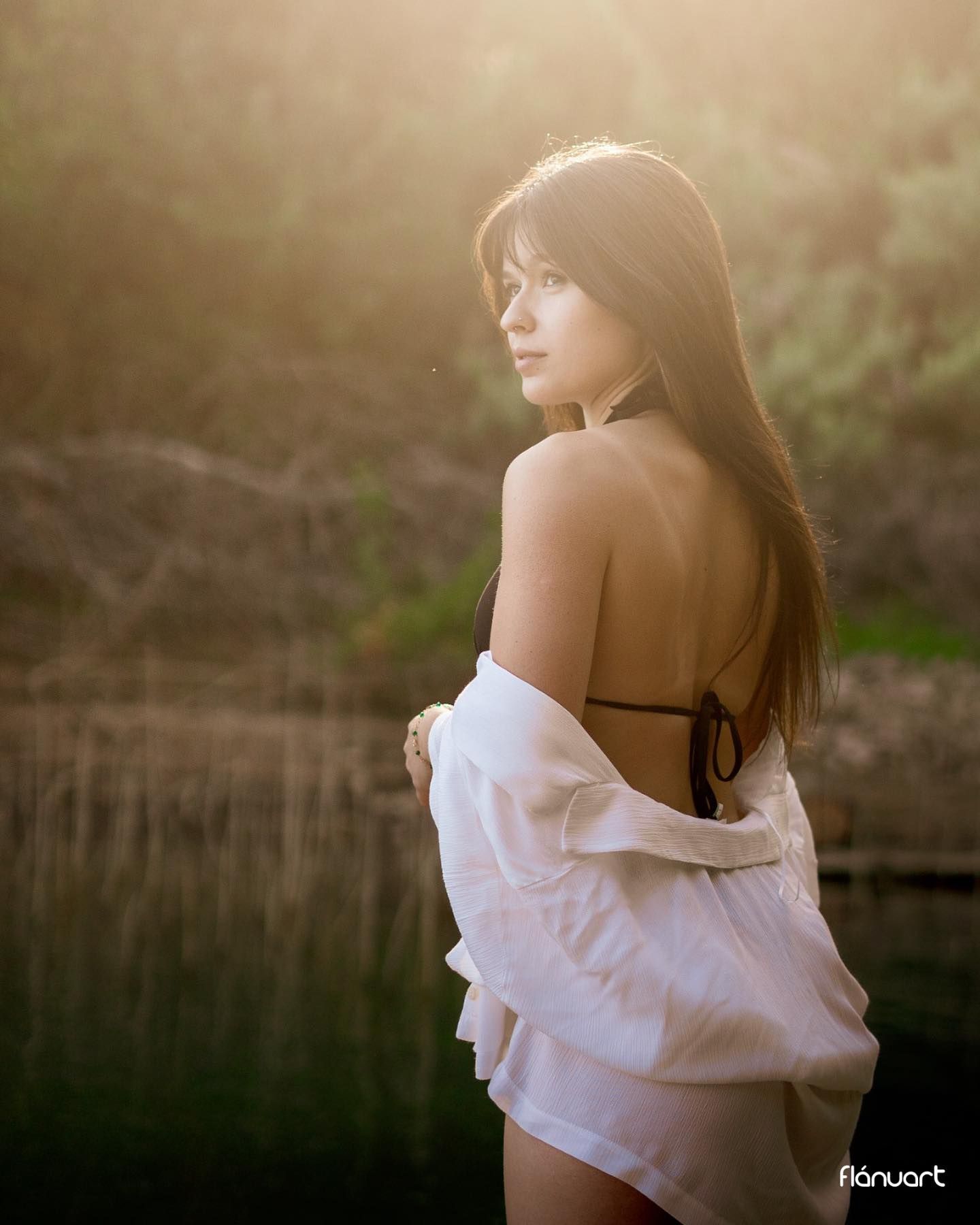 Woman in black swimsuit and open white shirt glowing in warm golden backlight by lake.