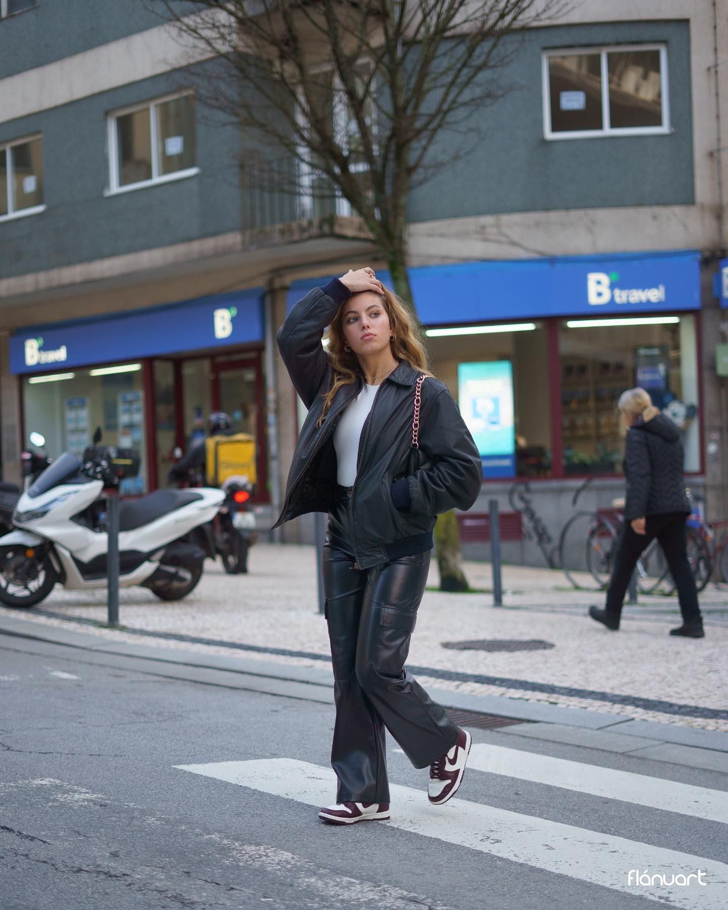 Stylish woman in black leather jacket and pants crossing a city street, wearing Nike sneakers.