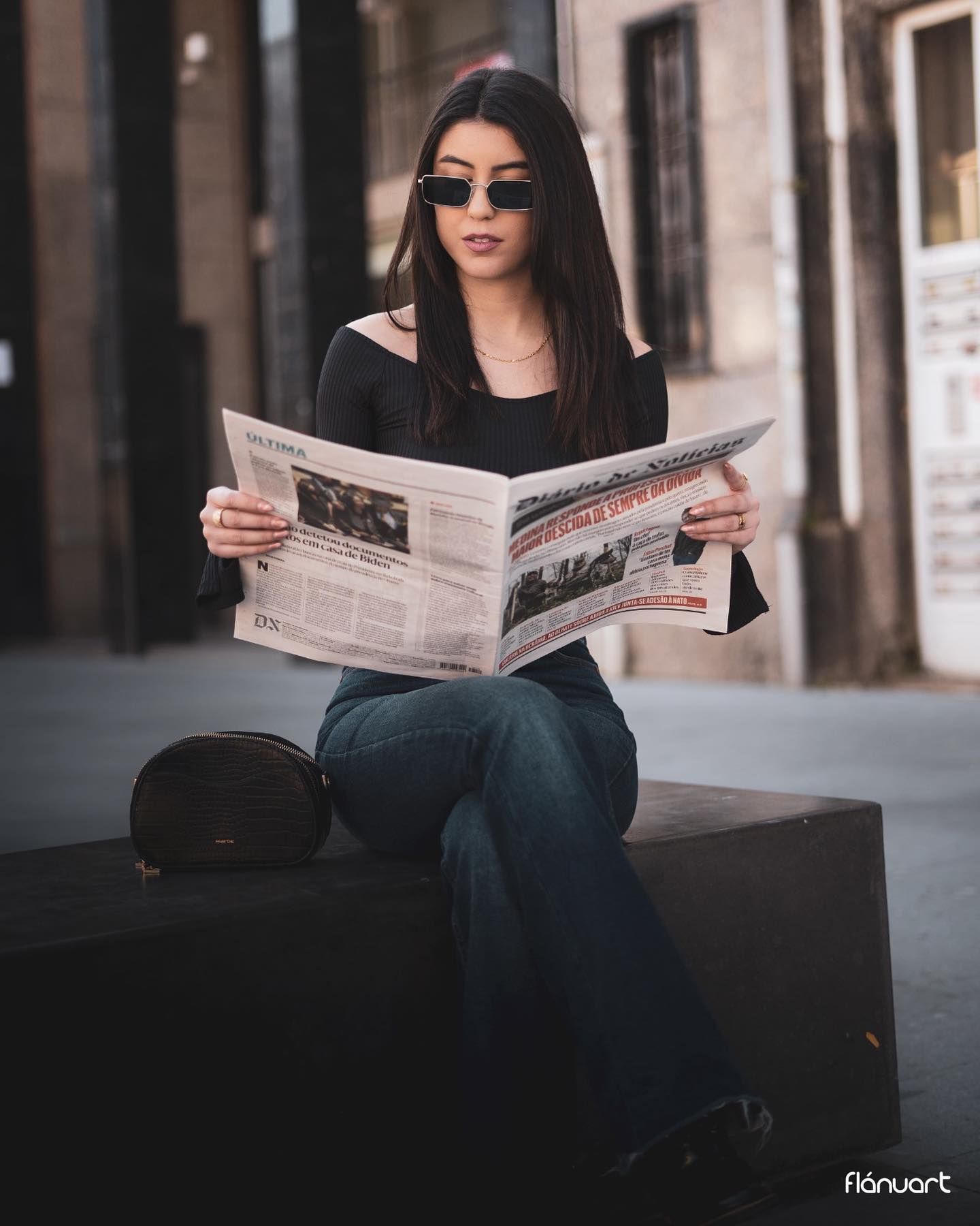 Young woman in black top and jeans reading newspaper outdoors, wearing sunglasses, seated on urban bench.