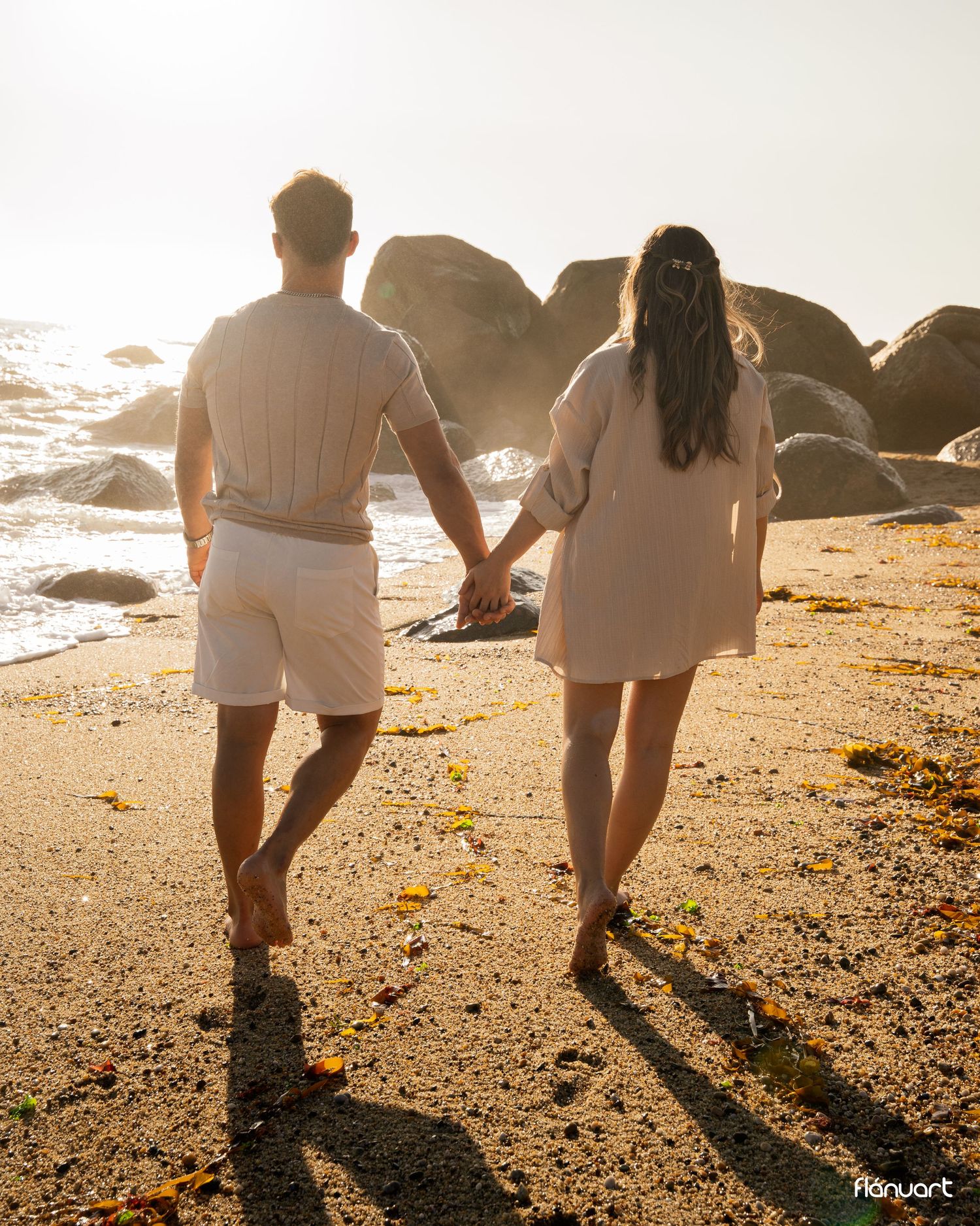 Couple holding hands walking barefoot on a sandy beach at golden sunset with rocks and ocean waves in the background.