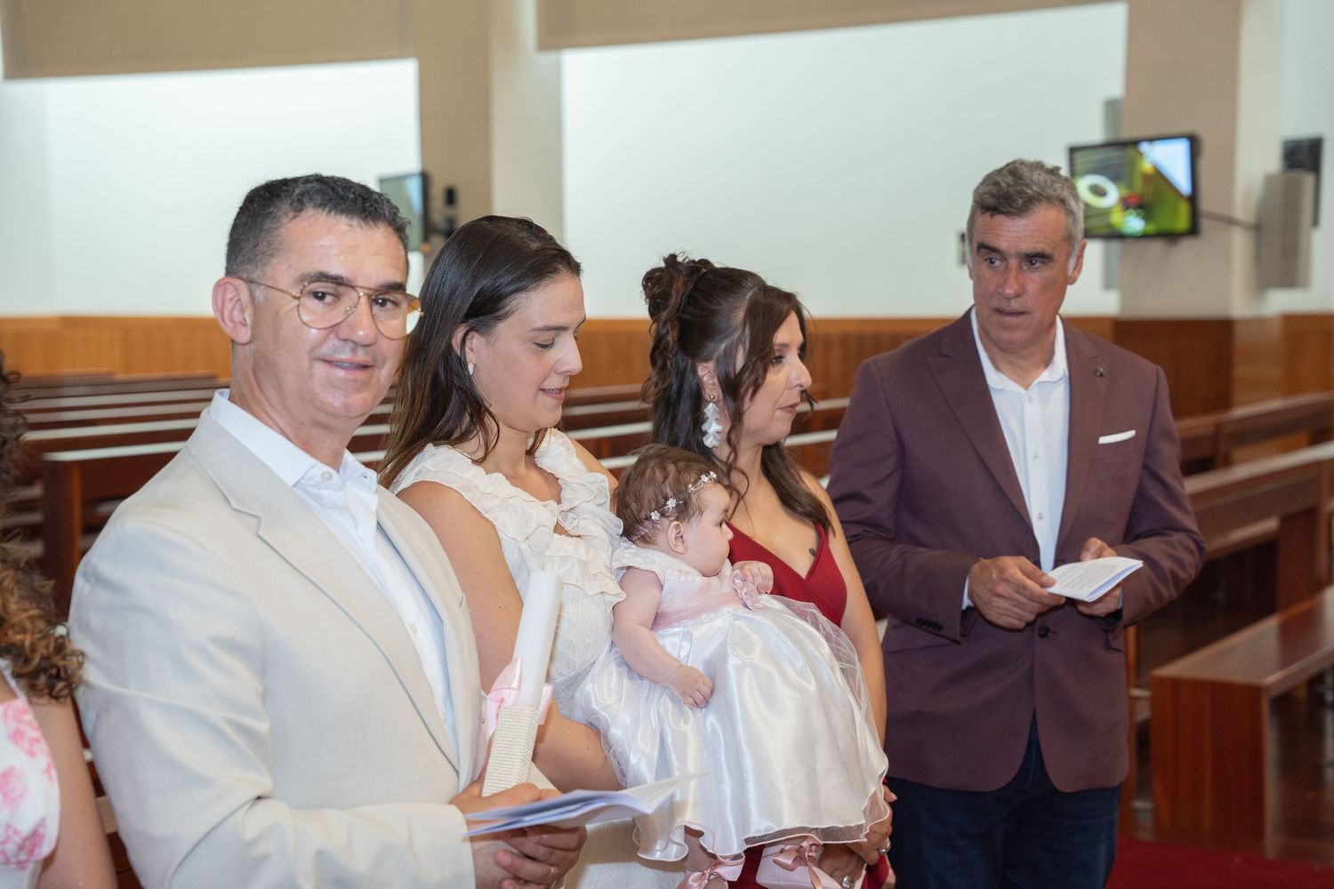 Family gathered in a church for a baptism ceremony, with parents holding a baby dressed in white christening gown.