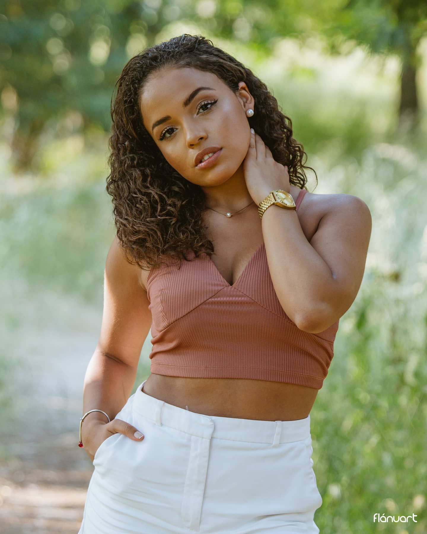 Young woman with curly hair in rust crop top and white skirt posing in park.