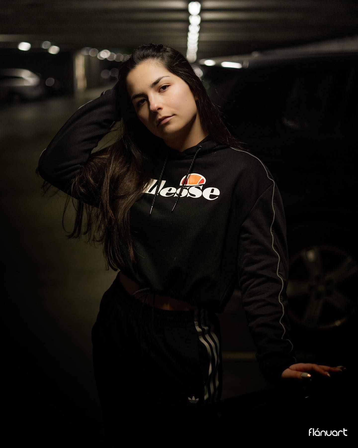 Young woman in black Ellesse hoodie and Adidas pants posing in a dimly lit parking garage.