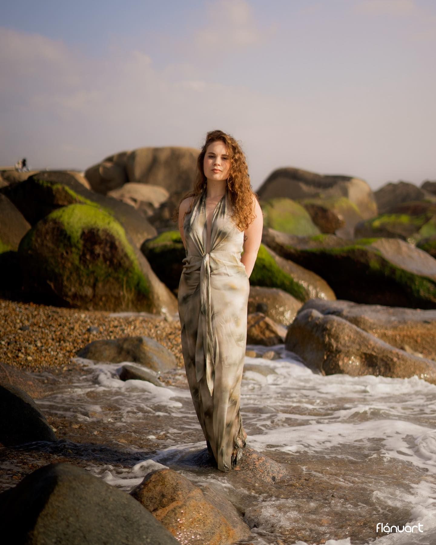 Redhead woman in elegant gray dress standing among mossy rocks on a rocky shoreline.