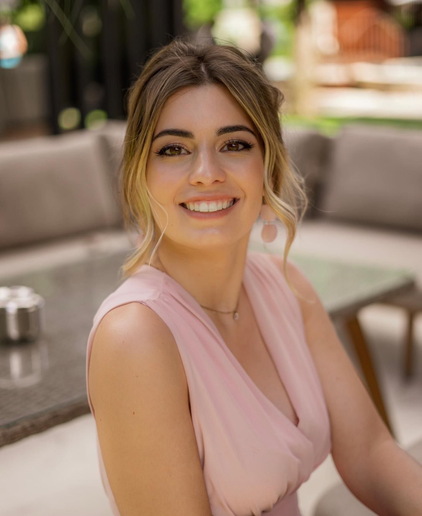 Smiling woman in pink dress at outdoor cafe with blurred background.