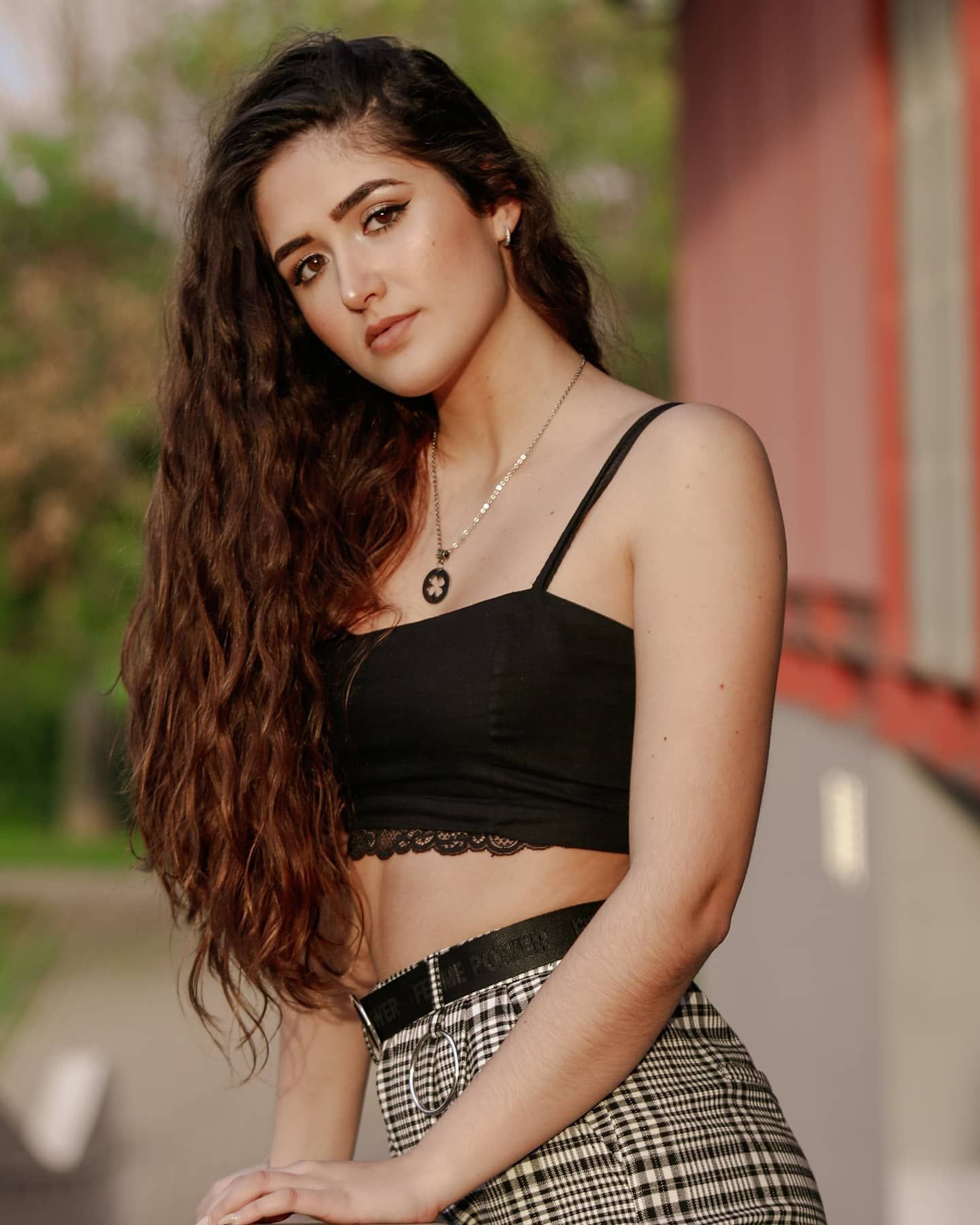 Brunette woman with long curly hair in a black lace crop top and plaid pants posing outdoors.