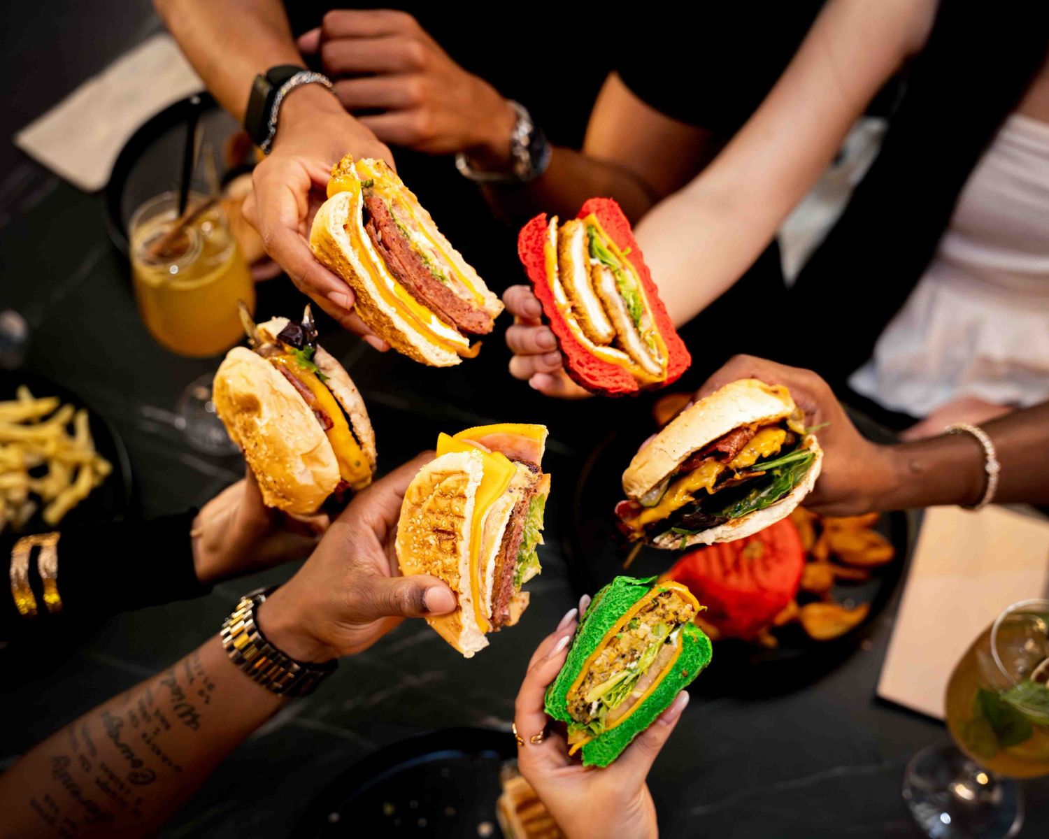 Friends sharing colorful gourmet burgers with vibrant buns at a restaurant table with drinks and fries.