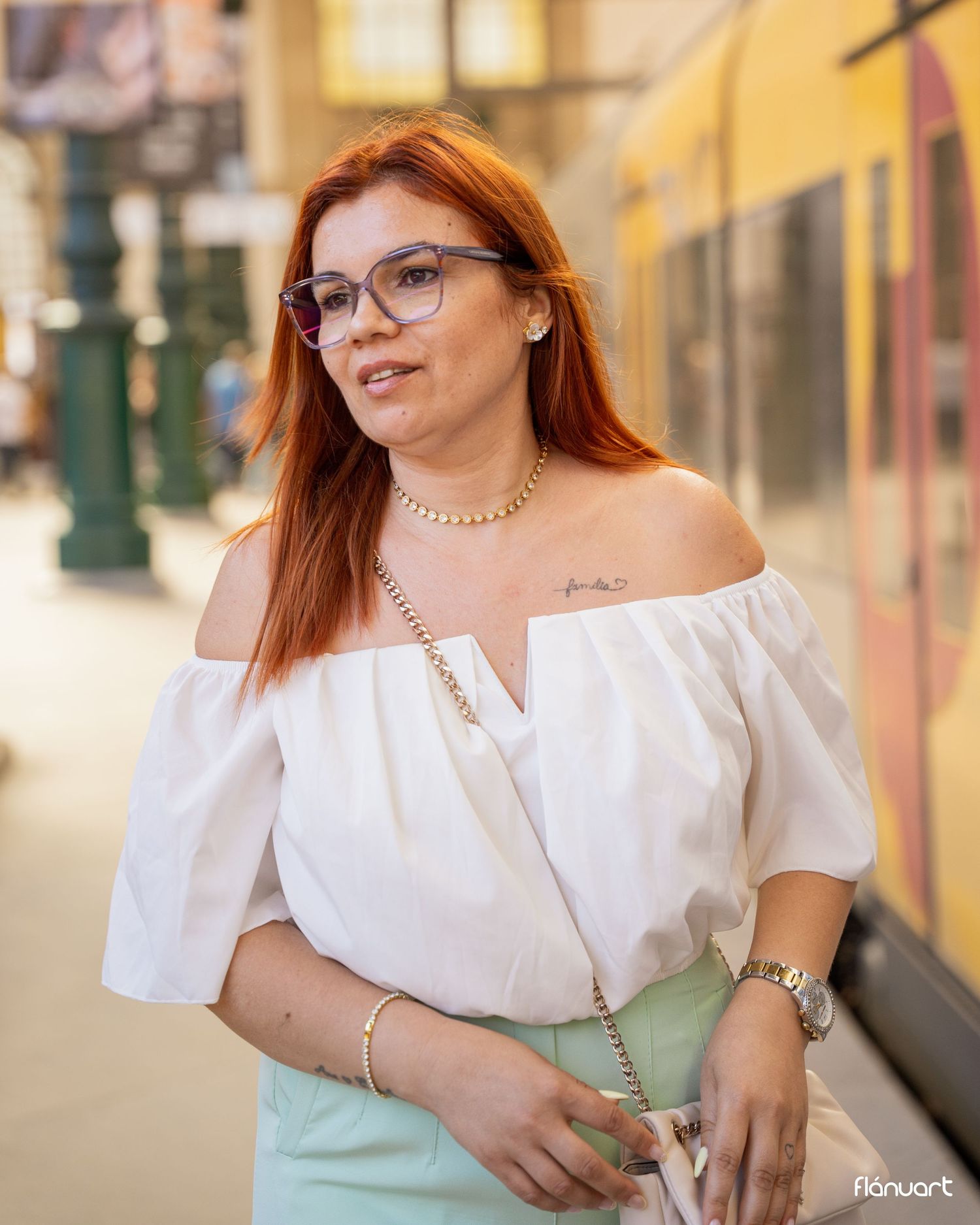 Stylish woman with red hair and glasses wearing white off-shoulder top and mint skirt at a city transit stop.
