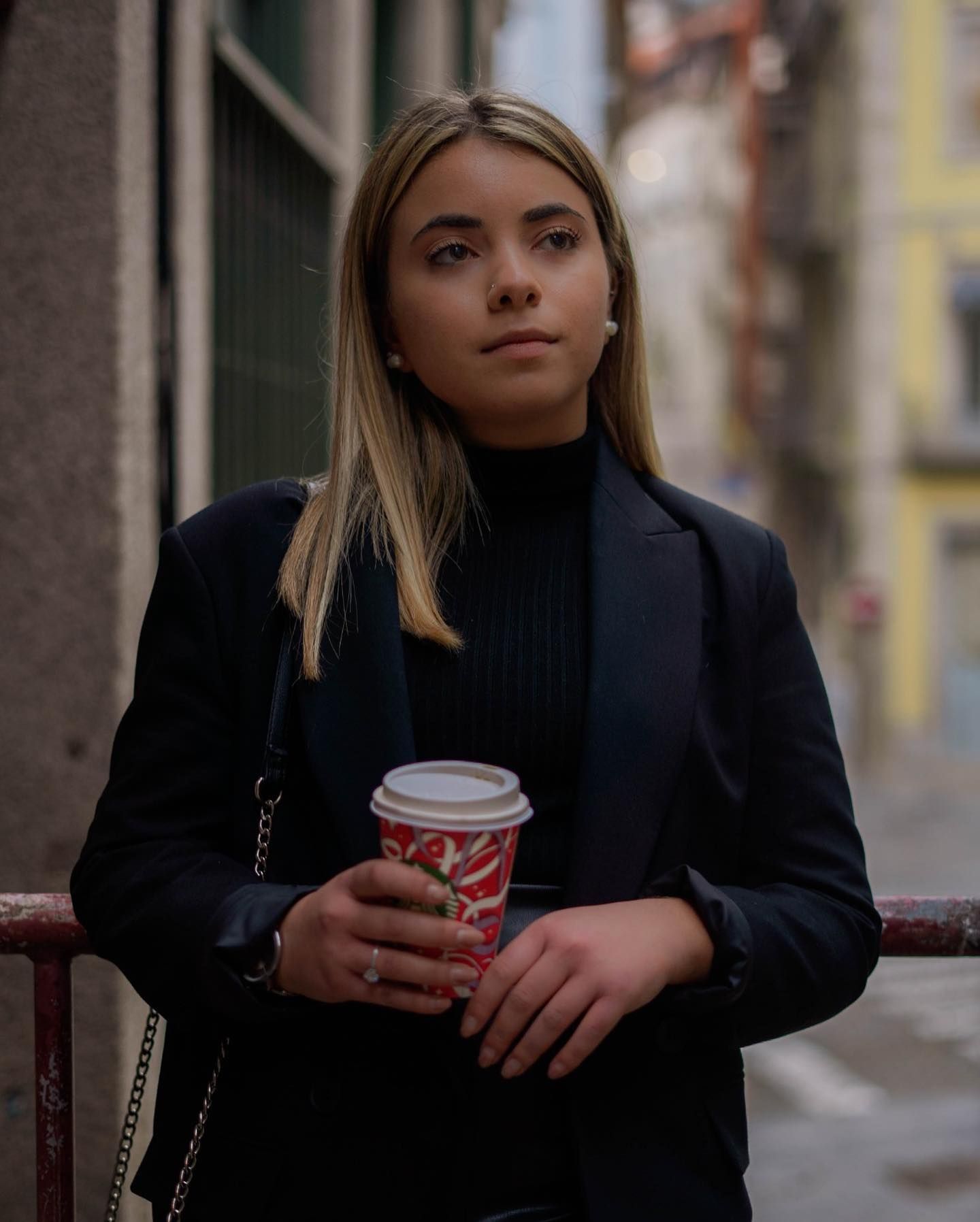 Blonde woman in black blazer and turtleneck holding a coffee cup on a city street.