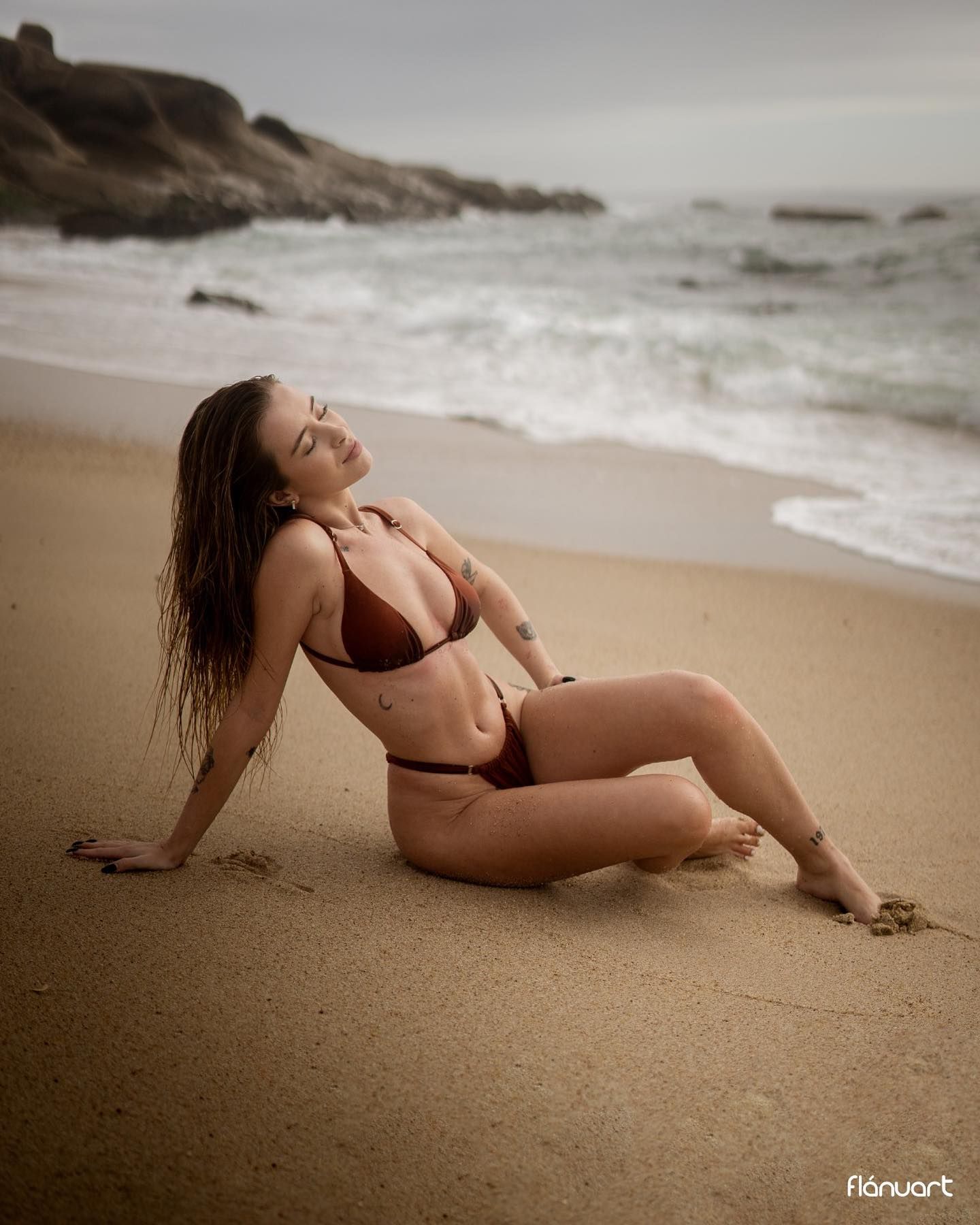 Woman in brown bikini relaxing on sandy beach with ocean waves behind her.