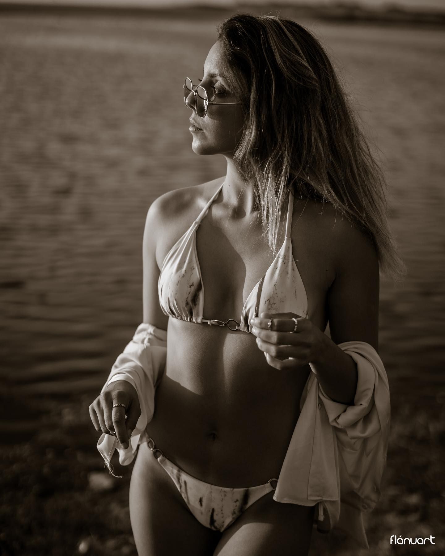 Sepia toned photo of woman in halter bikini and sunglasses posing by water.