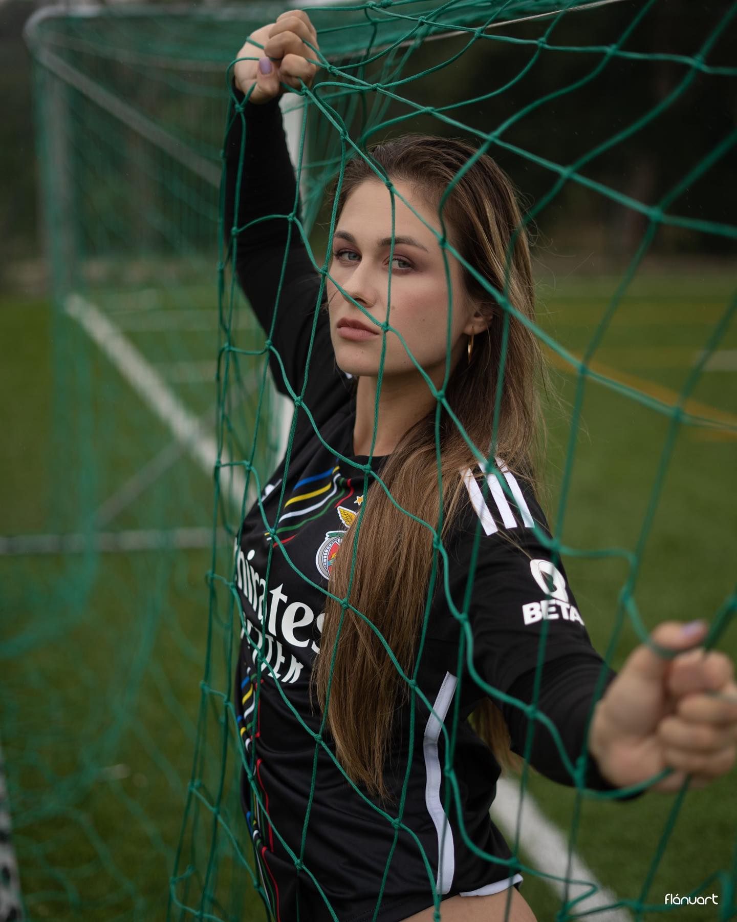 Young woman in black Adidas soccer jersey posing against a green goal net on a football field.
