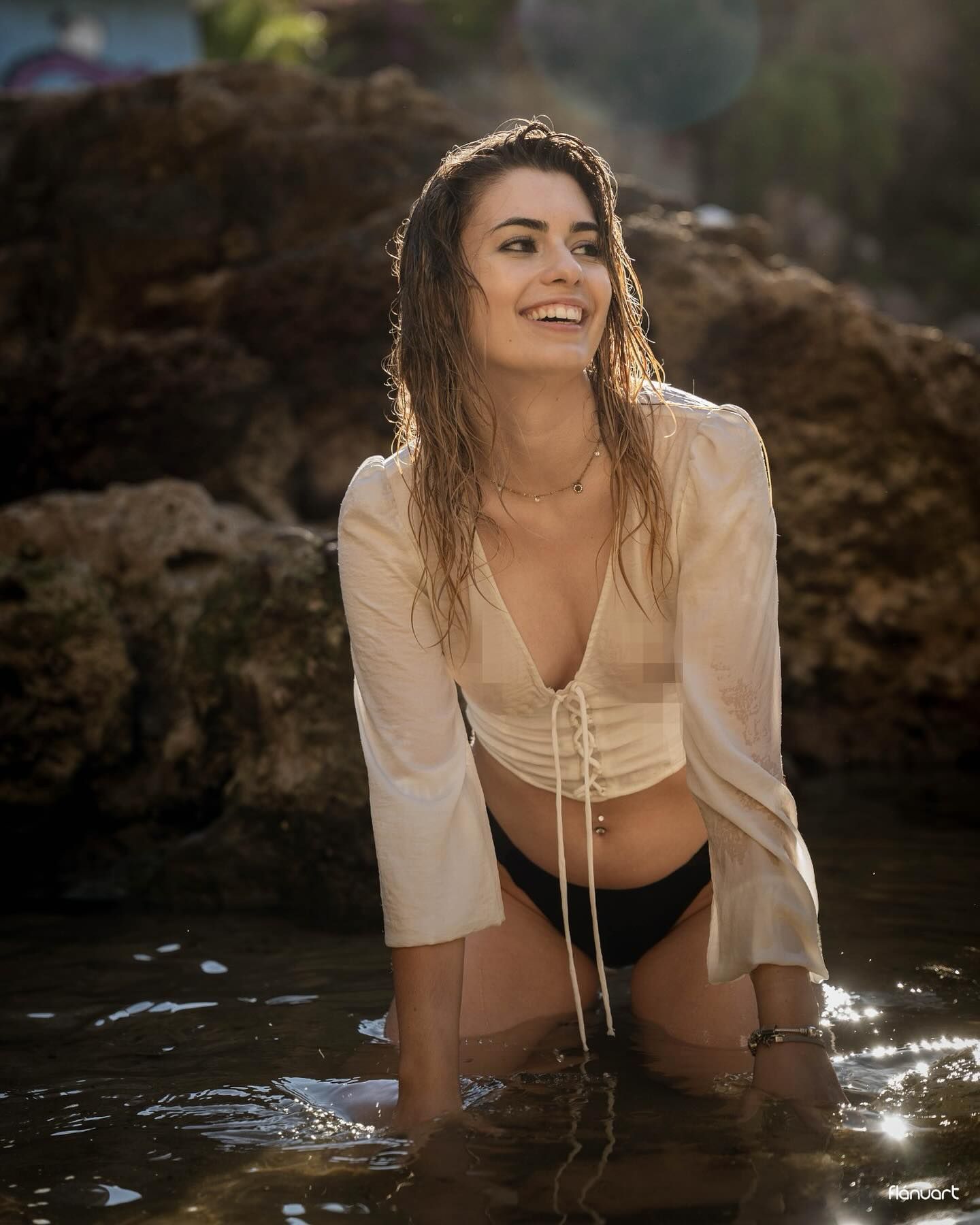 Smiling woman in white lace-up top and black bikini bottoms posing in shallow water near rocky shore.