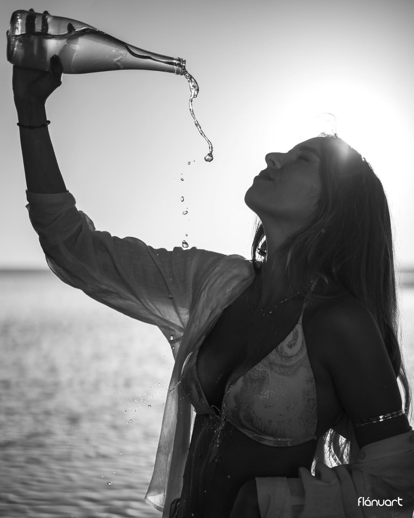 Black and white photo of woman pouring water from bottle overhead on sunny beach.