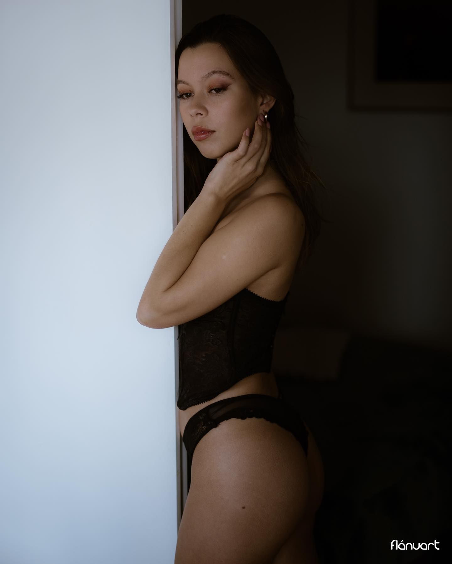 Woman in black lace bodysuit and thong posing beside a white wall in dim light.