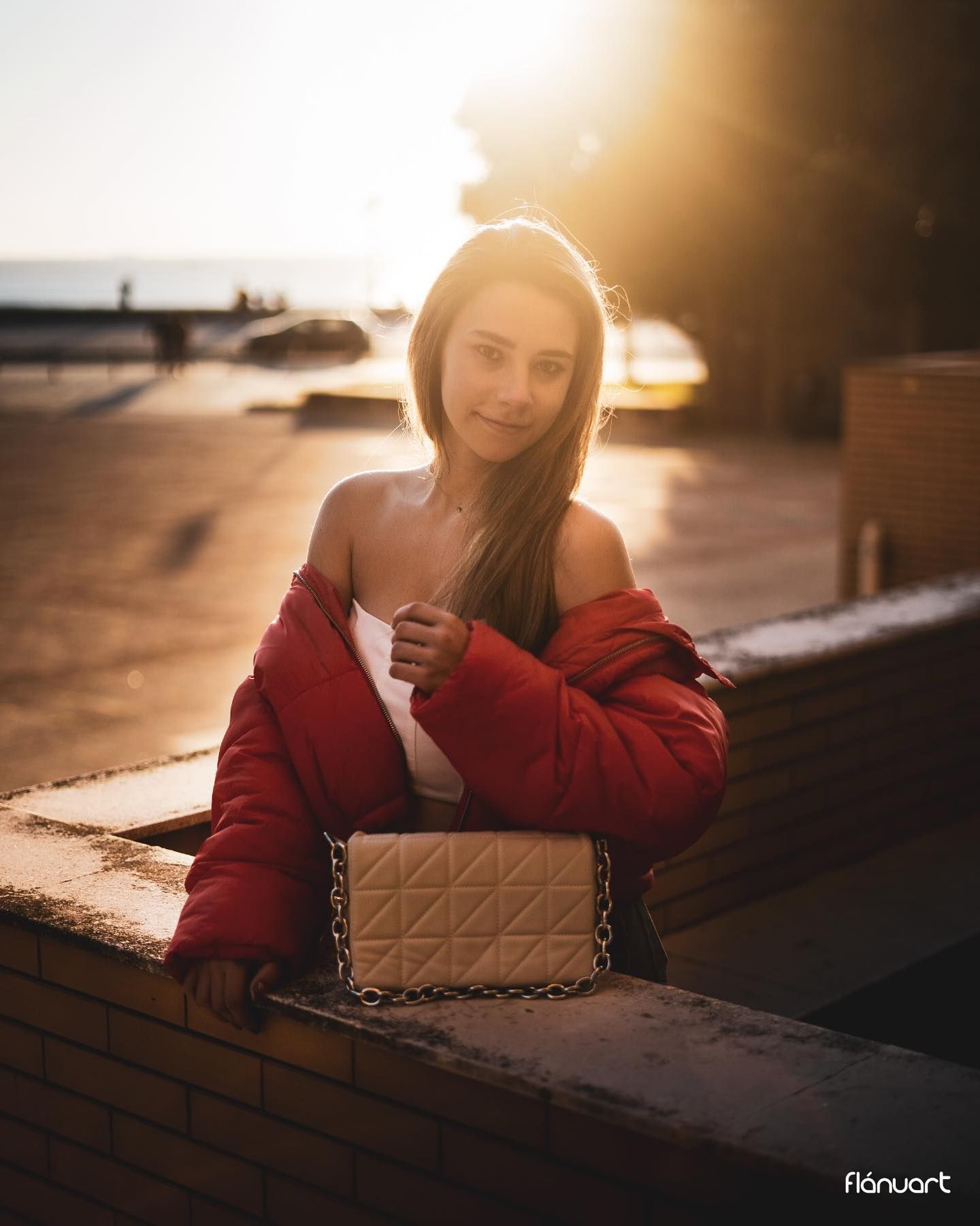 Young woman in a red jacket holding a beige quilted bag in golden sunset light outdoors.